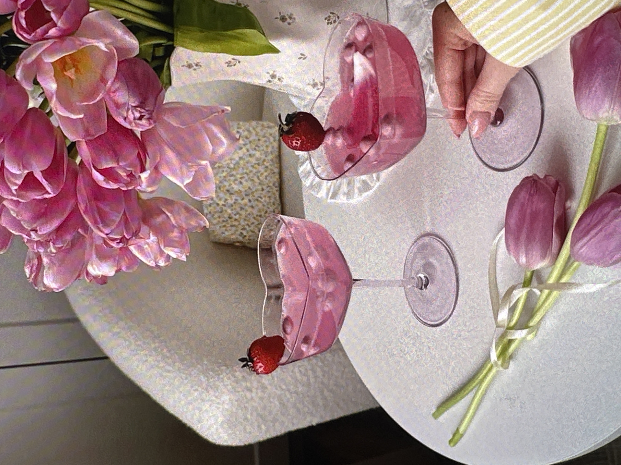 The image depicts a table setting, potentially in a cafe or residence in Yerevan, Armenia. On the table are two heart-shaped glasses filled with a pink liquid, each garnished with a strawberry. A bouquet of pink tulips sits on the left side of the table, while another bouquet of purple tulips, tied with a white ribbon, is placed to the right. A hand wearing a striped garment is touching one of the glasses. The table is covered with a light-colored cloth. A decorative box is visible in the background behind the glasses.