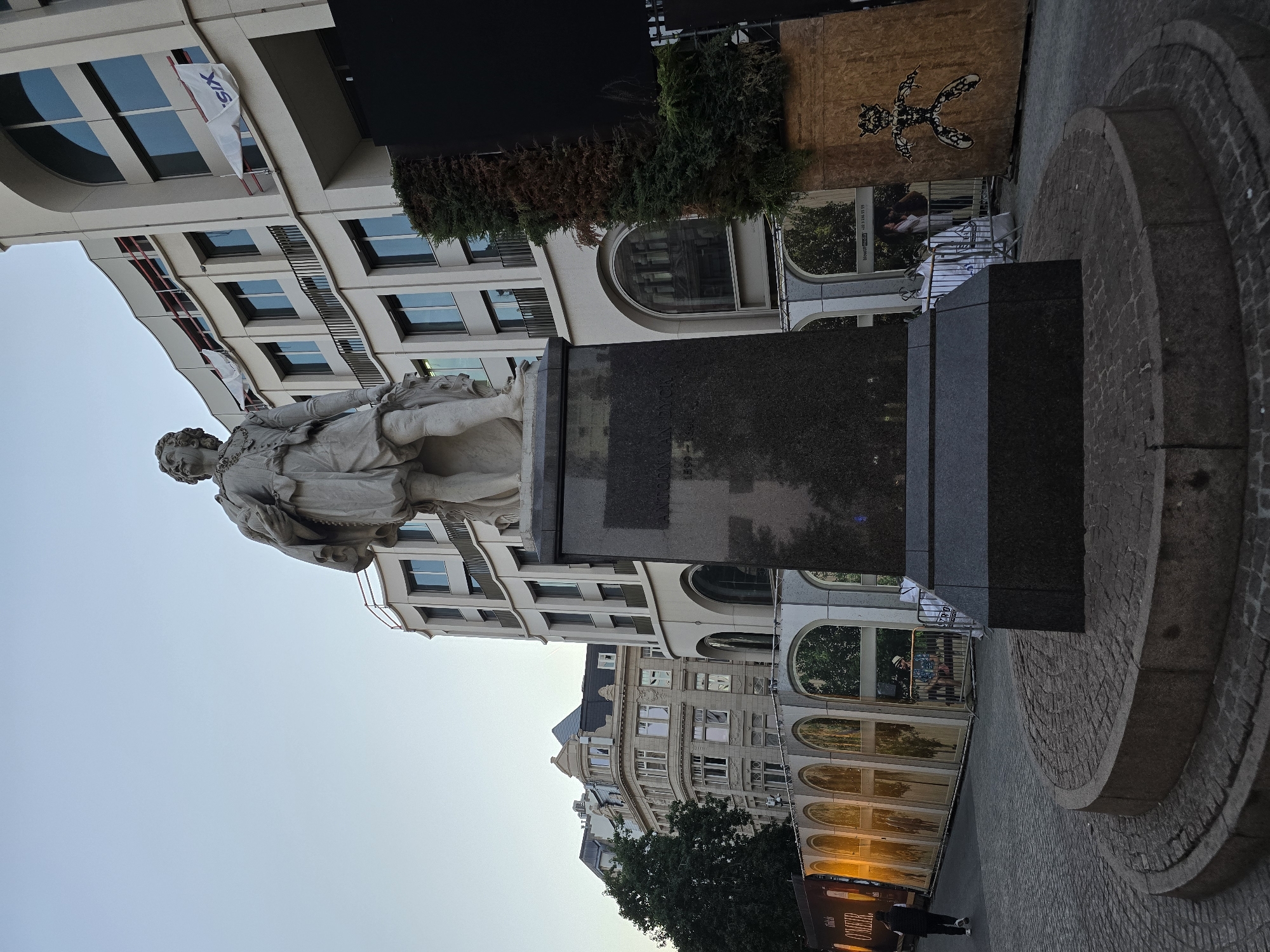 This is a low-angle, full shot of a statue of Peter Paul Rubens in the Grote Markt square in Antwerp, Belgium. The statue is made of white stone and depicts Rubens standing, holding a cloak and a piece of fabric. It stands on a tall, dark granite pedestal with some inscriptions visible. The statue is located in the foreground, with a cobblestone circular base. Behind the statue, there are several modern buildings with curved facades and arched windows. Some of the buildings have balconies. There are also some temporary structures, including a fence with people behind it, and a large wooden panel with a black stencil drawing of a figure. A banner with images of arches and trees is visible in the middle ground. The sky is clear and pale blue. The lighting suggests it is either early morning or late afternoon.