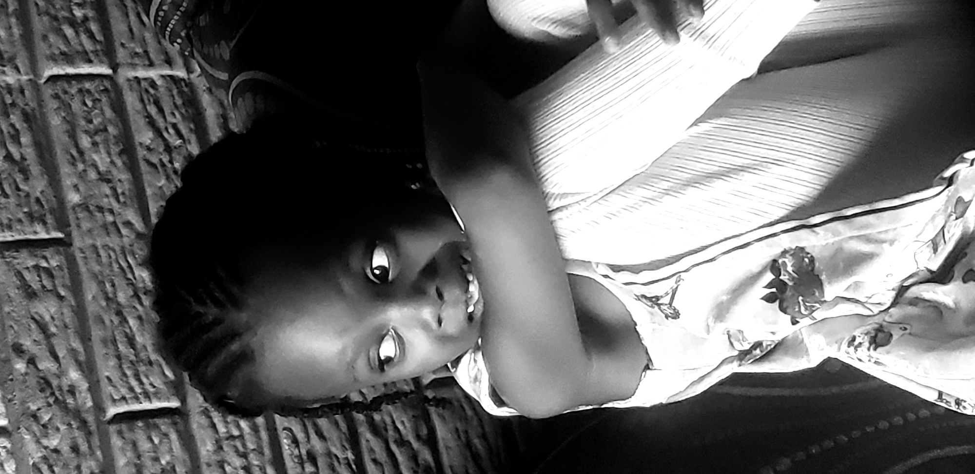 The black and white media file captures a young girl in a close-up shot, seemingly lying down. She has braided hair and is looking towards the camera with a slightly amused expression, her mouth open as if in laughter or speech. The background shows a textured surface, possibly a brick or stone wall, and what appears to be fabric with a floral pattern. The girl is wearing a top with thin straps and a ribbed texture on her arm. The lighting suggests an indoor setting, though the exact time of day is not discernible. The overall mood is candid and intimate. The location context is Narayi, Nigeria.