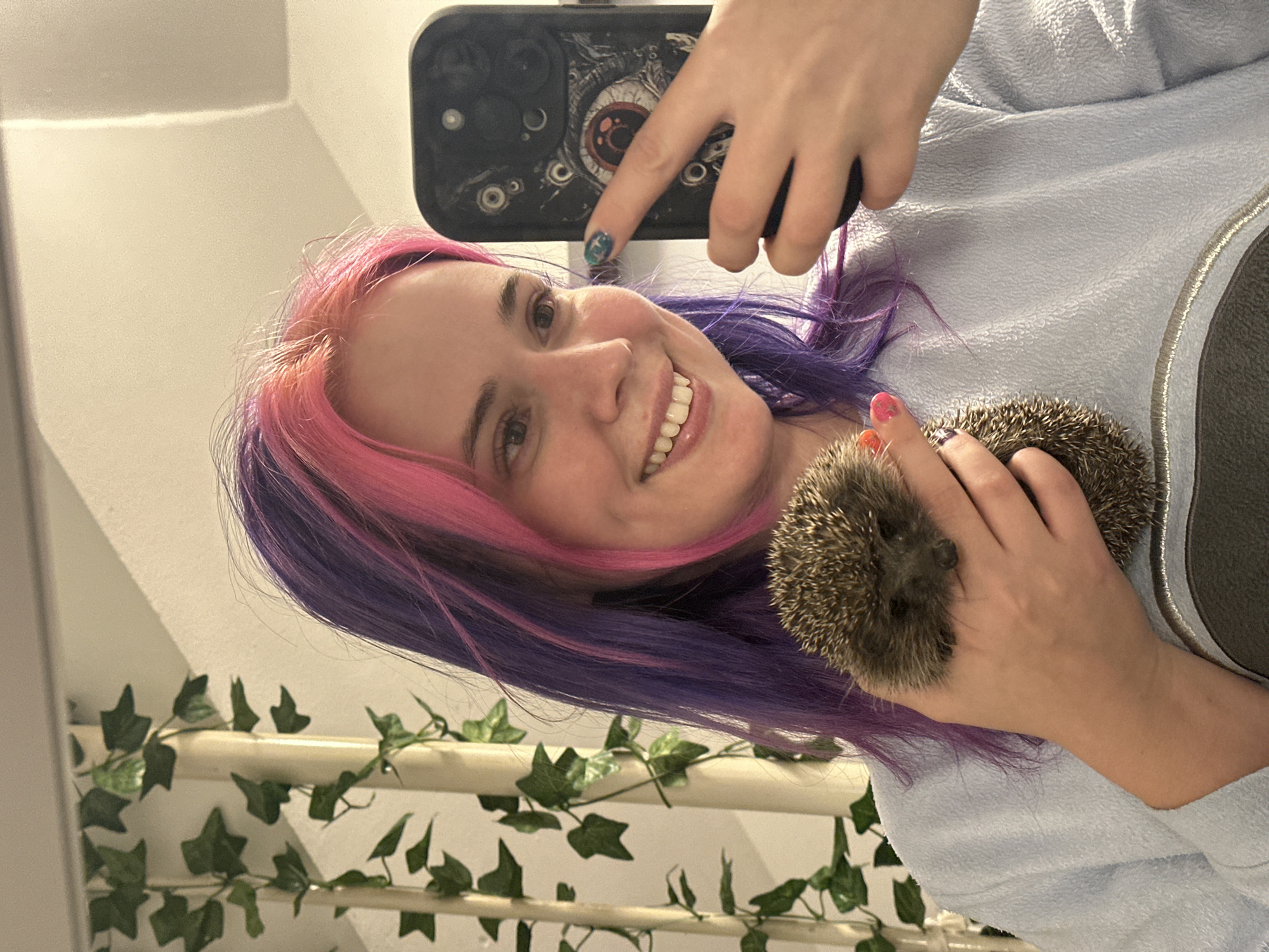 A woman with pink and purple hair is holding a hedgehog and taking a selfie. The phone has an eye-themed case. The background includes a staircase railing decorated with artificial ivy. The location appears to be indoors and is not explicitly identifiable as City of Zagreb, Croatia without additional context.