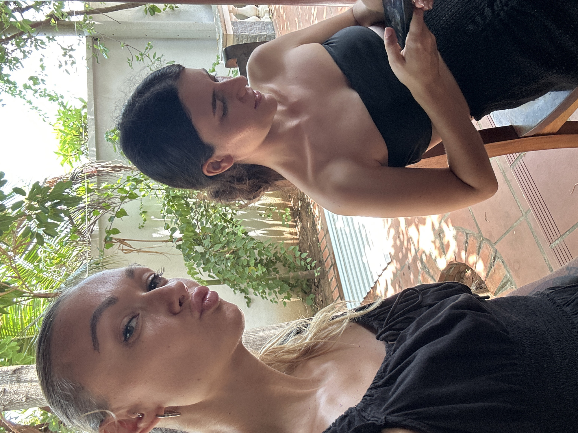 Two women are in the image. One has dark hair and is wearing a black strapless top. She appears to be looking at a phone. The other woman has blonde hair, pursed lips, and is wearing a black top. They appear to be outside in a location with plants and brick flooring. Given the location provided, this could be somewhere in Mũi Né, Vietnam, possibly a patio or garden area.
