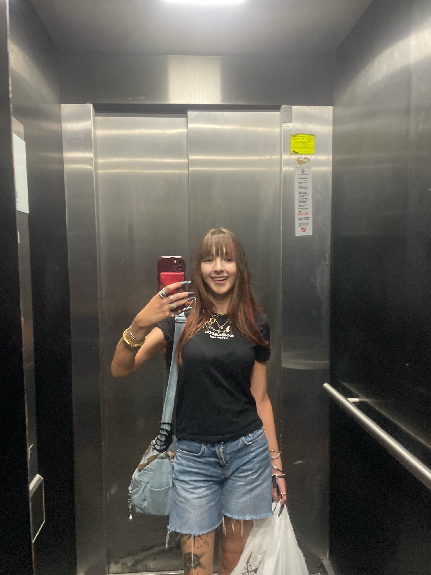 A young woman with long brown hair and bangs, wearing a black t-shirt and denim shorts, takes a selfie in an elevator. She is holding a red smartphone in her right hand and has several rings on her fingers and bracelets on her wrists. A light blue crossbody bag hangs over her left shoulder. She is smiling with her tongue sticking out. The elevator walls are made of brushed metal, and there are informational stickers on the right wall. The location is Raanana, Israel.