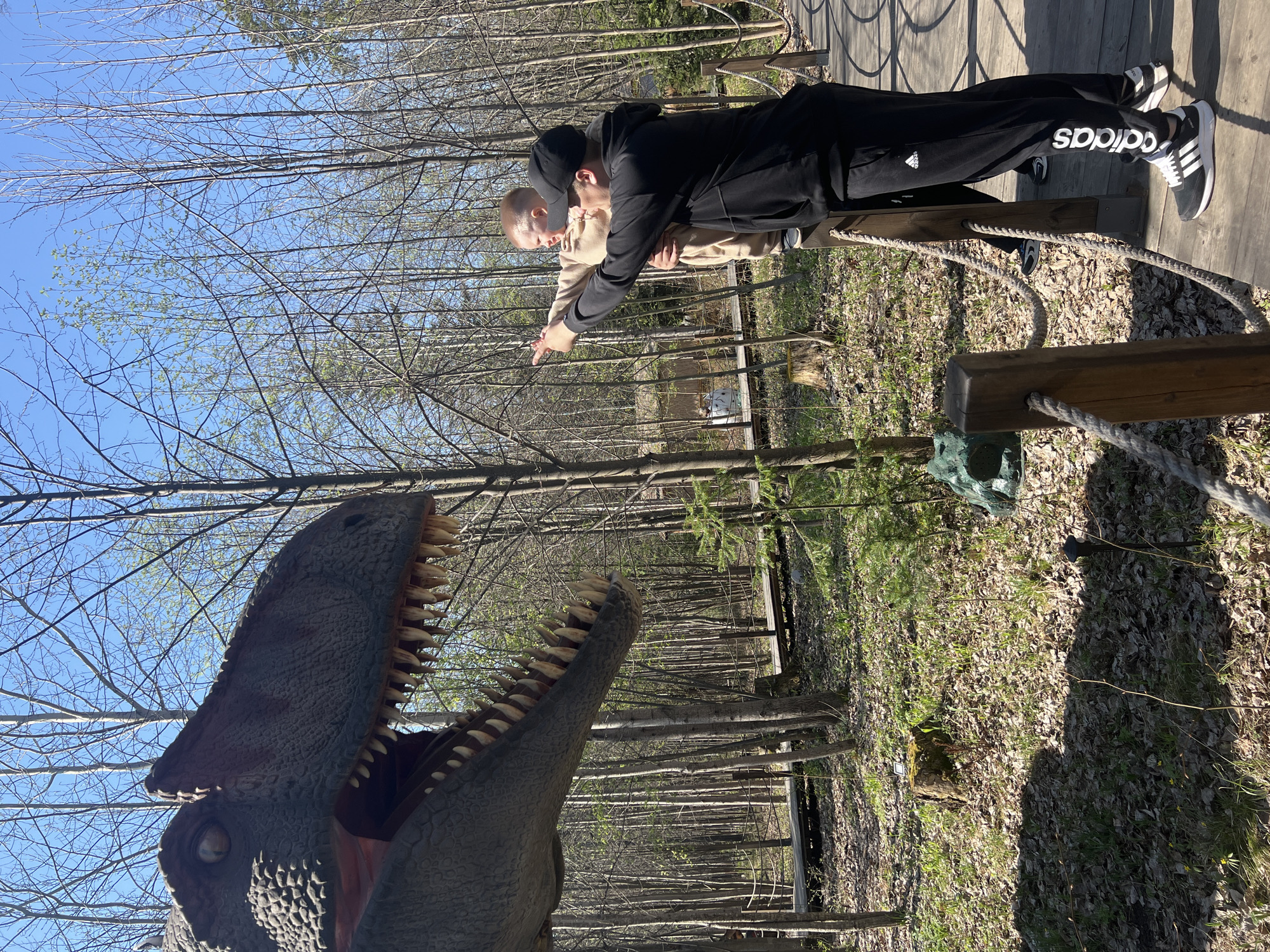 Two people are standing on a wooden walkway, pointing towards a dinosaur statue. One person is wearing a black hooded sweatshirt and black Adidas pants. The other person is wearing a light-colored shirt. The statue of the dinosaur has its mouth open, revealing its teeth. The location appears to be in a wooded area, with trees and foliage visible. The city of Izhevsk, Russia, is also identified as the location.