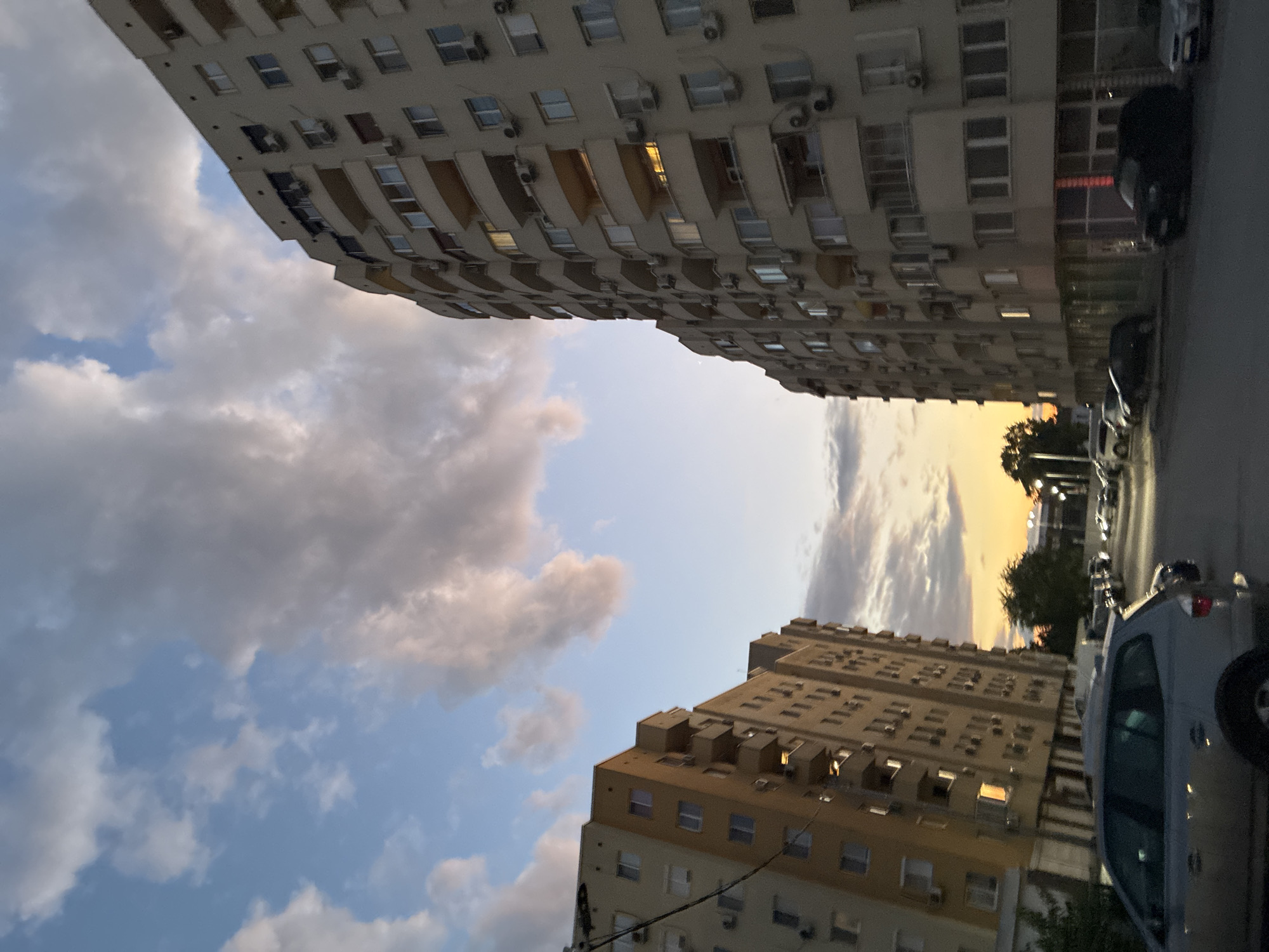 The image captures a street scene in Plovdiv, Bulgaria, during sunset. Two multi-story apartment buildings dominate the frame, one on the left with a yellow facade and balconies, and another on the right, a larger, curved building with a beige facade and numerous balconies. The sky above is a mix of blue and dramatic, colorful clouds, with the setting sun casting a warm glow on the horizon. A street with parked cars and a silver SUV in the foreground leads away from the viewer, between the two buildings. The architecture suggests a residential area typical of Soviet-era urban planning.