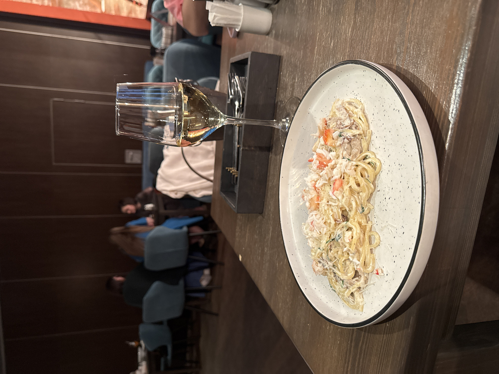 A plate of crab pasta and a glass of white wine sit on a wooden table at a restaurant in Moscow, Russia. Other patrons sit at tables in the background. A black utensil holder with silverware is next to the wine glass.