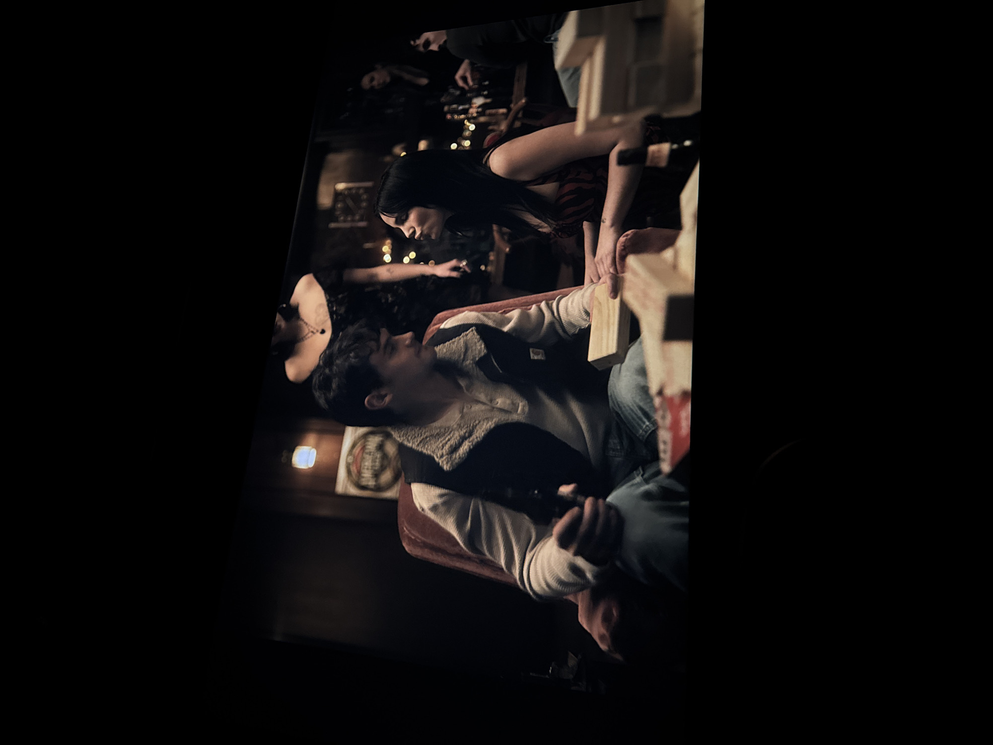 The image shows a dimly lit interior, likely a bar or pub, with several people gathered. A man in a light-colored shirt with a dark vest sits in a red chair, holding a beer bottle. A woman with long, dark hair leans towards him, her hands near a structure of wooden blocks. Other individuals are visible in the background, along with beer bottles and string lights. A sign with the word "AMERICANA" is visible on the wall. The location cannot be confirmed as Puebla, Mexico based on visual evidence.