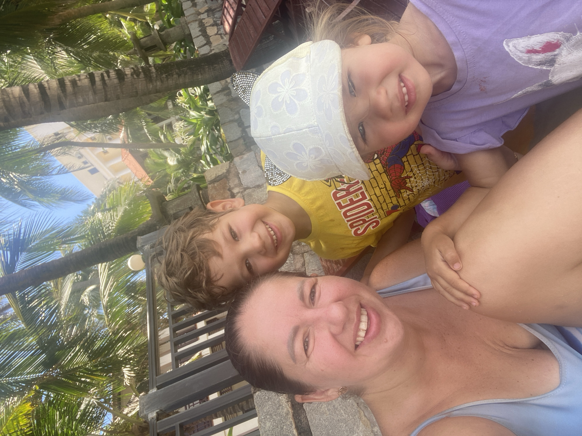 In a bright, outdoor setting possibly at a resort or beach in Phu Thuy Ward, Vietnam, a woman and two children are posing for a selfie. The woman, in the foreground on the left, is wearing a light blue swimsuit and smiling broadly. To her right is a young boy wearing a yellow t-shirt with a Spider-Man graphic. A younger child, wearing a white cap with floral patterns and a light purple t-shirt, is in the front right, making a face. The background features lush palm trees, a building with a light-colored facade, and some outdoor furniture. The lighting suggests it is daytime, likely sunny, contributing to a casual and vacation-like atmosphere.