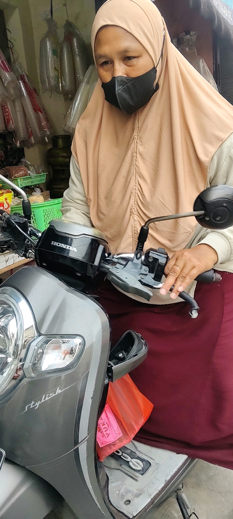 In Mekarmukti, Indonesia, a woman wearing a light brown hijab and a black face mask is seated on a grey scooter. She is wearing a light beige jacket over a maroon skirt. Her right hand is on the scooter's handlebar. In the background, shelves are stocked with items such as green baskets of goods, gas canisters, and plastic cups wrapped in plastic. An orange bag is tucked into the scooter's compartment in front of the woman.