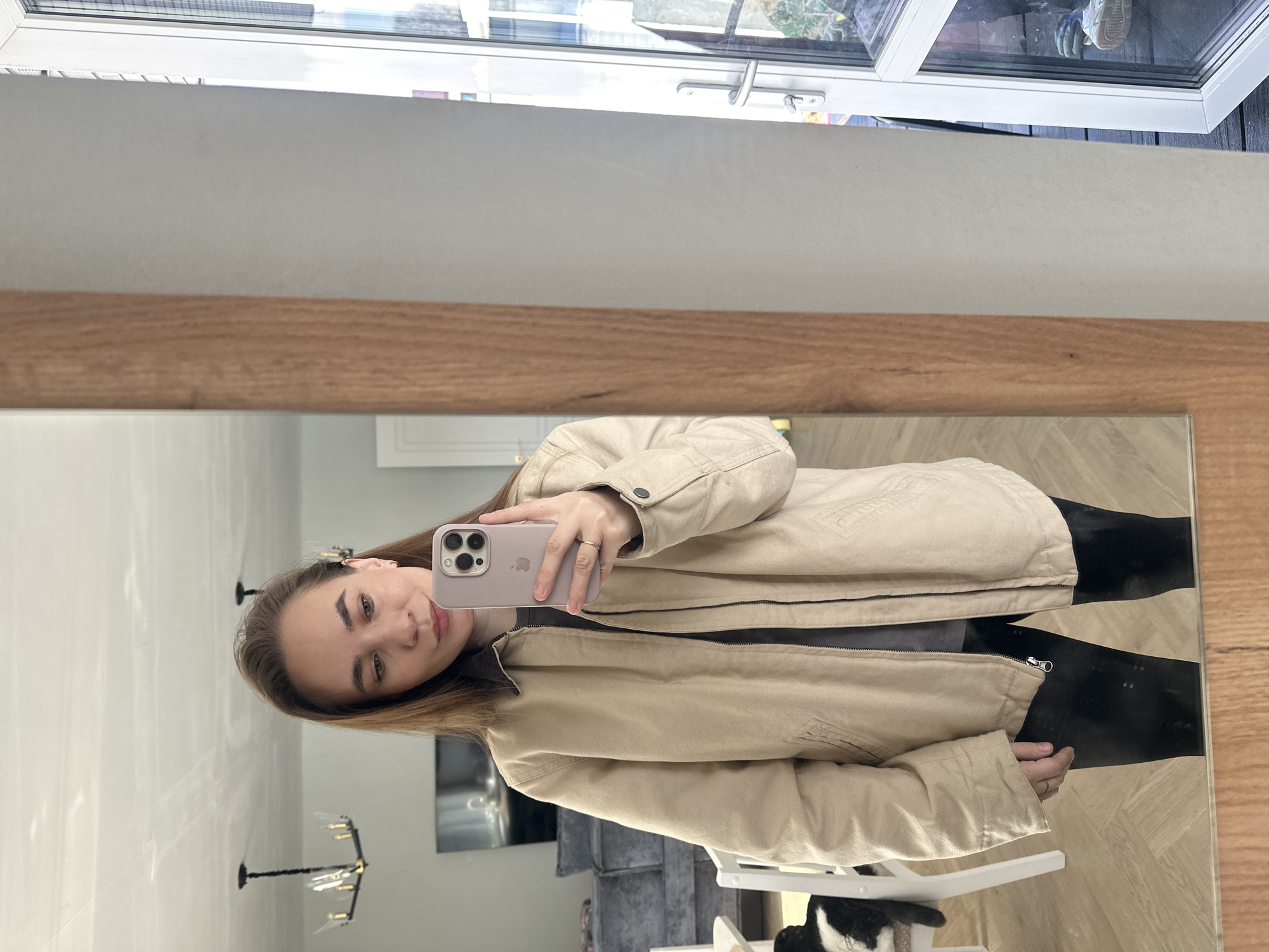 The image depicts an adult female in Kharnavy, Russia, captured in a mirror selfie within what appears to be a residential interior. The female, with long brown hair, is wearing a light beige zippered jacket with a dark brown collar, a dark grey shirt, and black leggings. She holds a light purple smartphone with a visible triple-lens camera module in her left hand, positioned to capture her reflection. A gold-colored ring is visible on her left ring finger.
The mirror's reflection reveals a contemporary living space. To the left, a grey sectional sofa and a large, dark flat-screen television are visible. Two black chandeliers with clear light shades are suspended from the ceiling. The flooring is light-colored, featuring a herringbone pattern. A white chair is partially visible in the lower left, upon which a black and white cat is seated. A white interior doorway is also reflected. To the right, the mirror's wooden frame abuts a light-colored wall, beyond which an open white-framed glass door or large window provides a view of an outdoor area with dark decking or patio surface, some foliage, and a silver-colored door handle.
