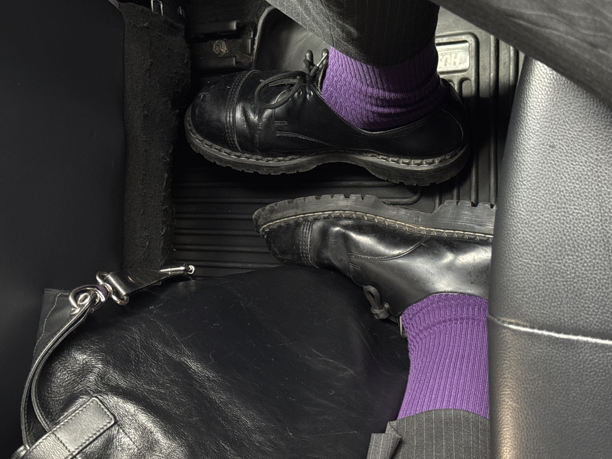 The image shows a portion of a car interior with a person's feet and legs. The person is wearing dark pinstripe pants and purple socks. The feet are shod in black leather shoes with thick soles. One foot is on the car floor near a black leather bag with silver hardware, while the other is near the gas pedal. The environment appears to be the inside of a car, with leather seats and textured floor mats. There is no indication of the city of New York.