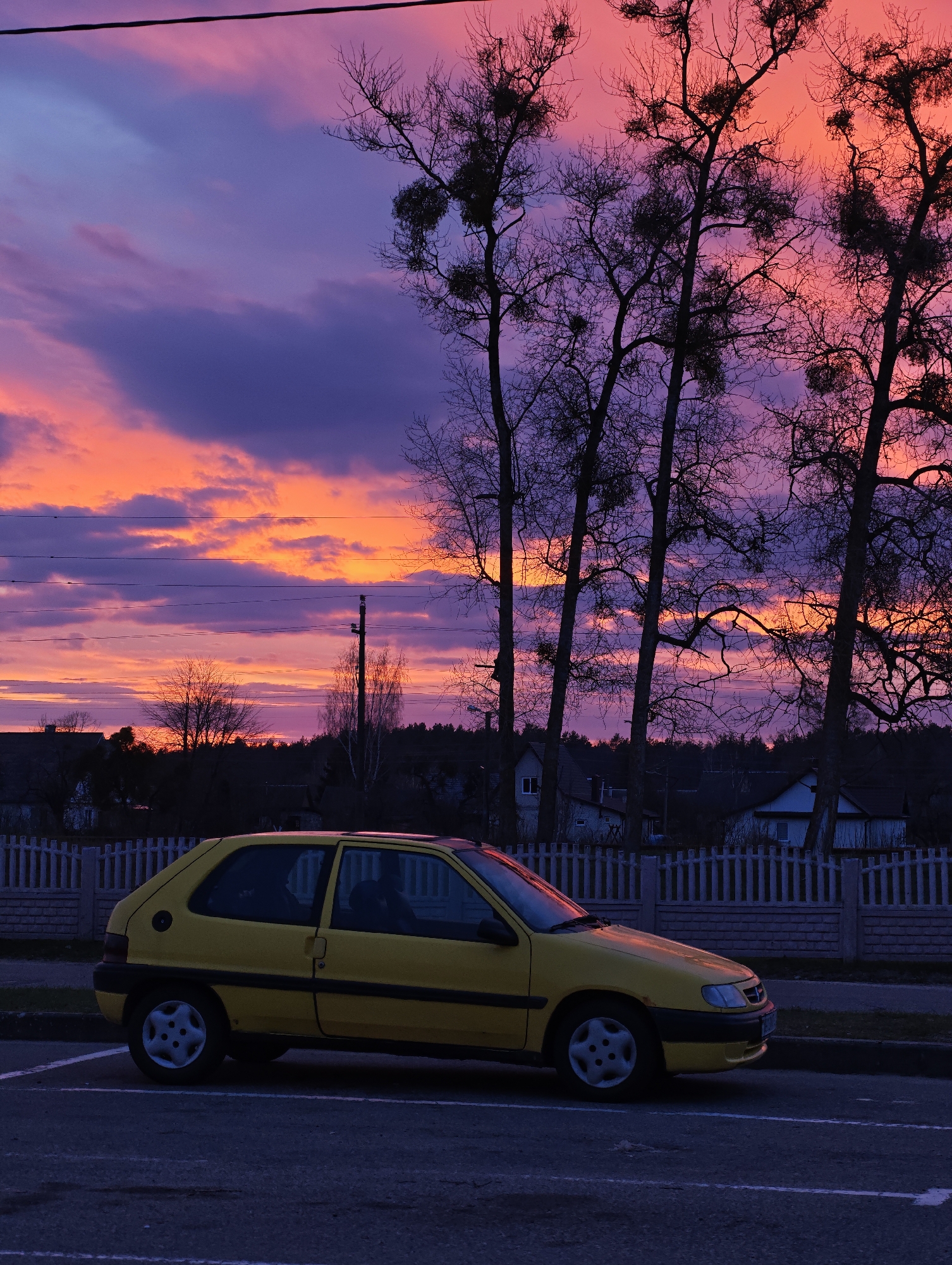 A yellow Citroen Saxo is parked in a parking space alongside a gray picket fence. Behind the fence are houses, and further back, a line of trees obscures the horizon. The sky is a dramatic mix of pink, orange, and purple hues, likely during sunset or sunrise. Three bare trees stand to the right of the car, their branches silhouetted against the colorful sky. Power lines are visible against the sky in the background. The location is a residential area, presumably in the city of Unknown, Unknown.