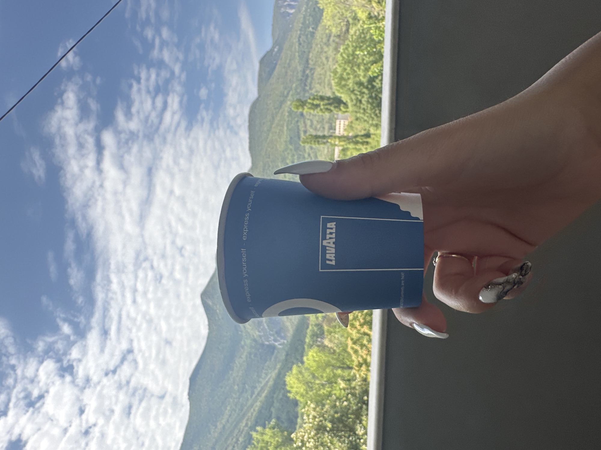 A woman's hand holds a blue Lavazza coffee cup against a backdrop of green mountains and a blue sky with scattered white clouds. The woman has white, almond-shaped fingernails with some having a marbled design. She is also wearing a small gold ring on her ring finger. The coffee cup is partially obscuring the view of the landscape. The overall scene suggests a person enjoying a moment of relaxation outdoors, likely on a balcony or terrace, with a scenic view. The image does not provide any specific details to identify the location as Vratsa, Bulgaria.