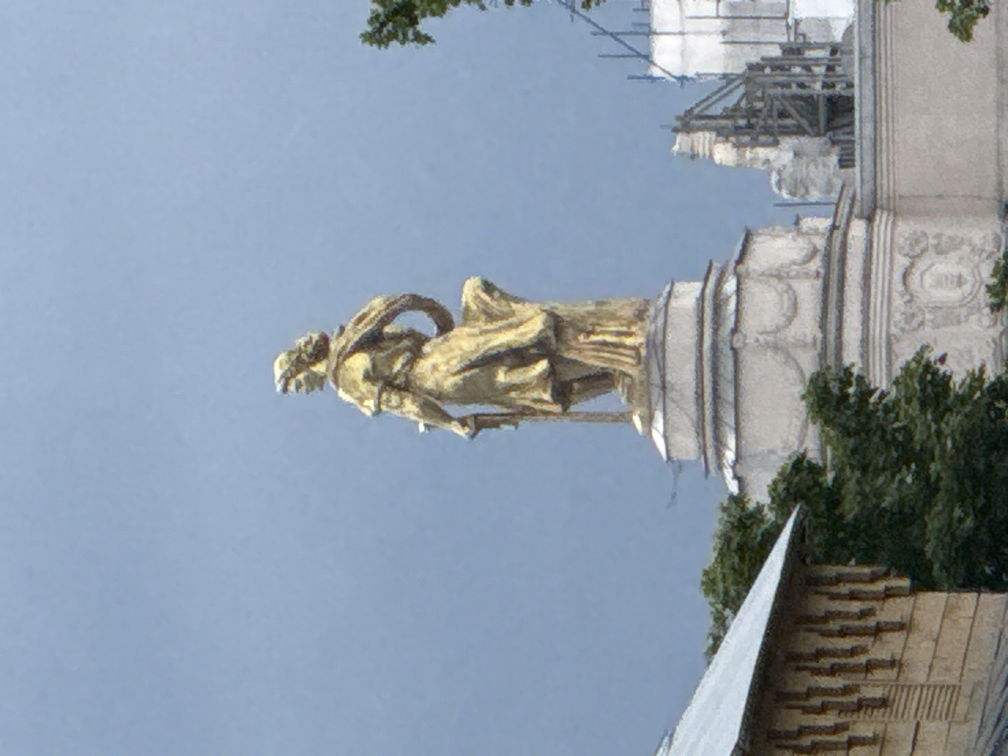 A golden statue of a female figure stands prominently against a pale blue sky, on top of a multi-tiered white pedestal. The statue depicts a woman in classical attire, with her left arm bent and her hand resting on her hip, while her right arm is extended downwards, holding a sword. She is facing to the right of the viewer. In the background, a portion of a building with a gray, sloped roof and wooden siding is visible on the left. To the right of the statue, parts of other buildings are visible, including a light-colored structure with decorative elements and some scaffolding or construction work. Green trees are interspersed in the mid-ground. The overall impression is of a historical monument in an urban setting. This statue is located in Moscow, Russia.