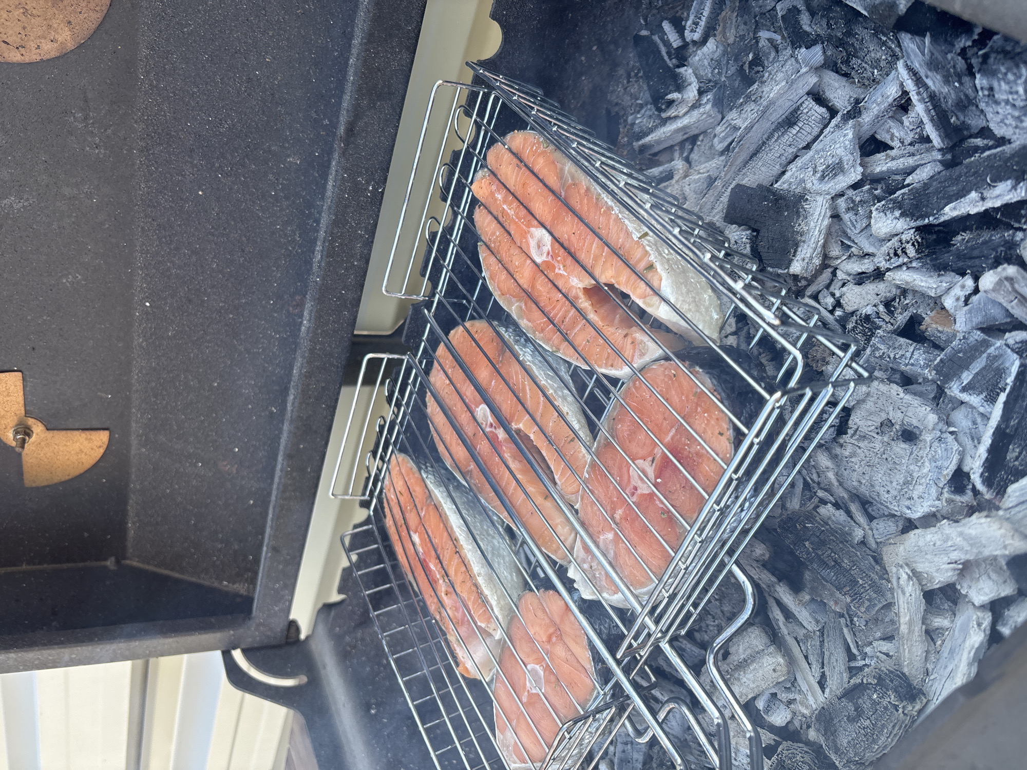 The image depicts pieces of raw salmon being grilled over hot coals in a barbecue. The fish is placed in a wire grilling basket. The grill appears to be a domestic barbecue grill, and the coals are grey and black. The location is inferred to be Chaikovsky, Russia, based on the prompt, but the image provides no concrete evidence to confirm this.