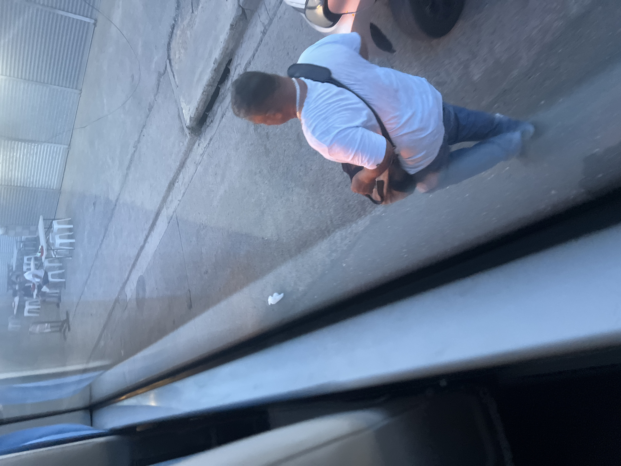 The image depicts a man in a white t-shirt and blue jeans walking away from the camera on a concrete surface. He is wearing a black strap across his shoulder, carrying a brown bag. In the background, there are several white tables and chairs set up, suggesting an outdoor dining or seating area. A corrugated metal wall runs along the back of the scene. A dark colored vehicle is partially visible on the right side of the image. The lighting suggests it is either early morning or late afternoon. The presence of outdoor seating and the general ambiance are consistent with casual dining establishments found in Davao City, Philippines, particularly in areas with an outdoor market or street food scene.