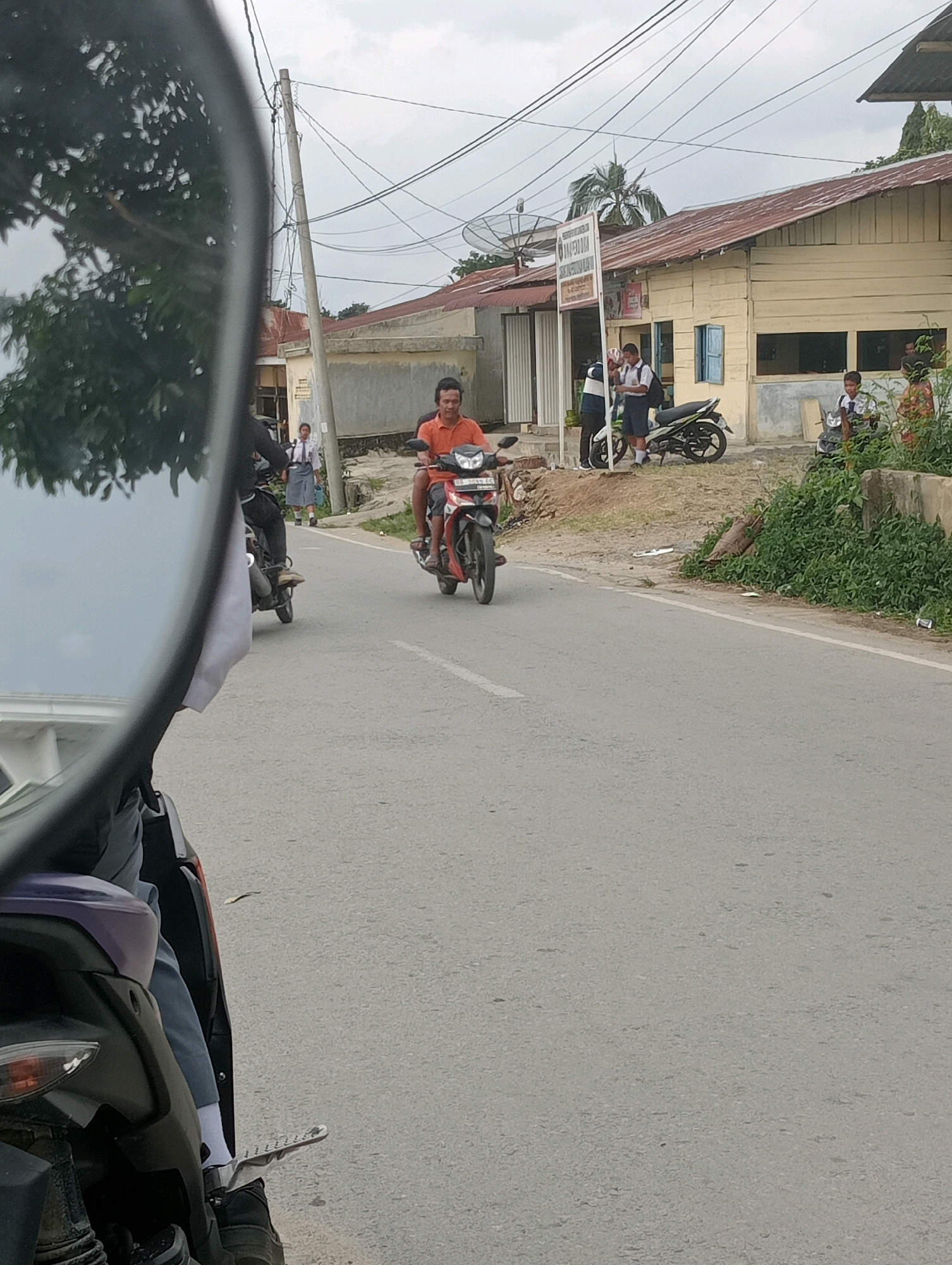 The image depicts a street scene in Hinalangbagasan, Indonesia, with a focus on the road and its surroundings. In the foreground, a motorcycle viewed from the rear is partially visible, with its rider's legs and feet clad in dark shoes and white socks. The rider is wearing dark pants. The road itself is asphalted and appears to be a two-lane street.
In the middle ground, a man is riding a red motorcycle towards the camera. He is wearing an orange t-shirt and red shorts. Several other motorcycles are parked or in motion in the background. A group of young people in school uniforms, likely students, are standing near a building. One student is on a white and green motorcycle. Another student is standing on the side of the road.
The buildings in the background are single-story structures with light-colored walls and red roofs. Some of the buildings have windows and doors. A signpost with writing is visible in front of one of the buildings. A satellite dish is mounted on the roof of a building. The environment is somewhat overgrown with green vegetation along the side of the road and near the buildings. The sky is overcast. The overall impression is of a typical day in a community with prevalent motorcycle use.