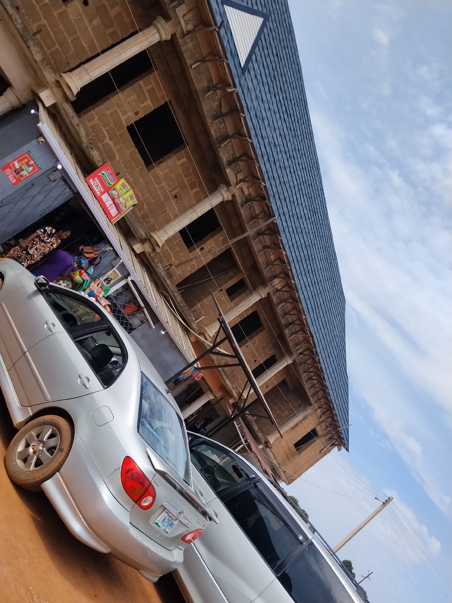 The image captures a street scene in Nabatum, Nigeria. A light-colored sedan, possibly a Toyota Corolla, is parked in the foreground, facing left. The car exhibits a sporty rear bumper with a spoiler and has a license plate that reads "IJN 258MB". Beside it, another vehicle, a white van or SUV, is partially visible.
Behind the vehicles, a two-story building is under construction. The lower level features an open storefront with merchandise displayed. Two individuals are visible near the storefront; one person, wearing a patterned dress, stands near the entrance, while another, in a purple garment, is further inside. Several colorful items, possibly toys or household goods, are arranged on shelves.
The building's facade is constructed from light brown bricks, with decorative columns supporting the upper level. The upper story has large, dark openings that appear to be windows or doorways. The building is topped with a dark blue, textured roof, and a white triangular section is visible near the roofline.
The sky above is partly cloudy, with a mix of blue sky and white clouds. Electrical wires are strung between poles in the background, indicating the presence of infrastructure. The ground appears to be dirt or unpaved road.