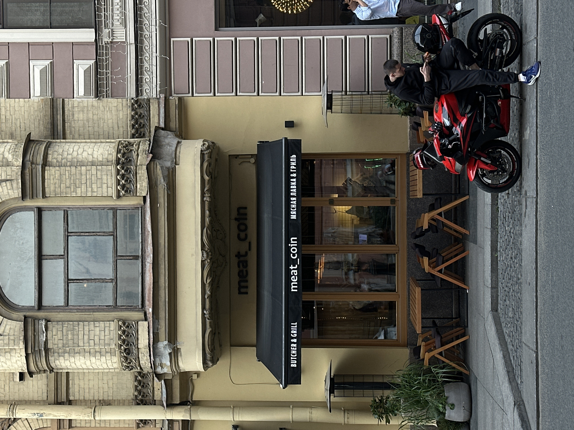 The image depicts a street scene in Saint Petersburg, Russia, outside a restaurant named "Meat_coin". The restaurant has a black awning with white lettering that reads "BUTCHER & GRILL" on the left and "meat_coin" and Russian text on the right. Beneath the awning, large glass windows reveal the interior, where mannequins dressed in clothing are displayed. Wooden tables and chairs are set up outside the restaurant, suggesting an outdoor seating area.
In the foreground, a red and black motorcycle is parked on the sidewalk. A young man wearing a black hoodie and black track pants is leaning against the motorcycle, looking down at his mobile phone. He is wearing blue and white sneakers. To the right of the motorcycle, another man in a light blue checkered shirt is standing and looking towards the right.
The architecture of the building is notable, with ornate brickwork and decorative elements above the restaurant. A large, arched window is visible on the upper floor. The street is paved with cobblestones and asphalt. Lush greenery is present in a large planter box next to the restaurant entrance. The overall atmosphere suggests a casual urban setting in Saint Petersburg.