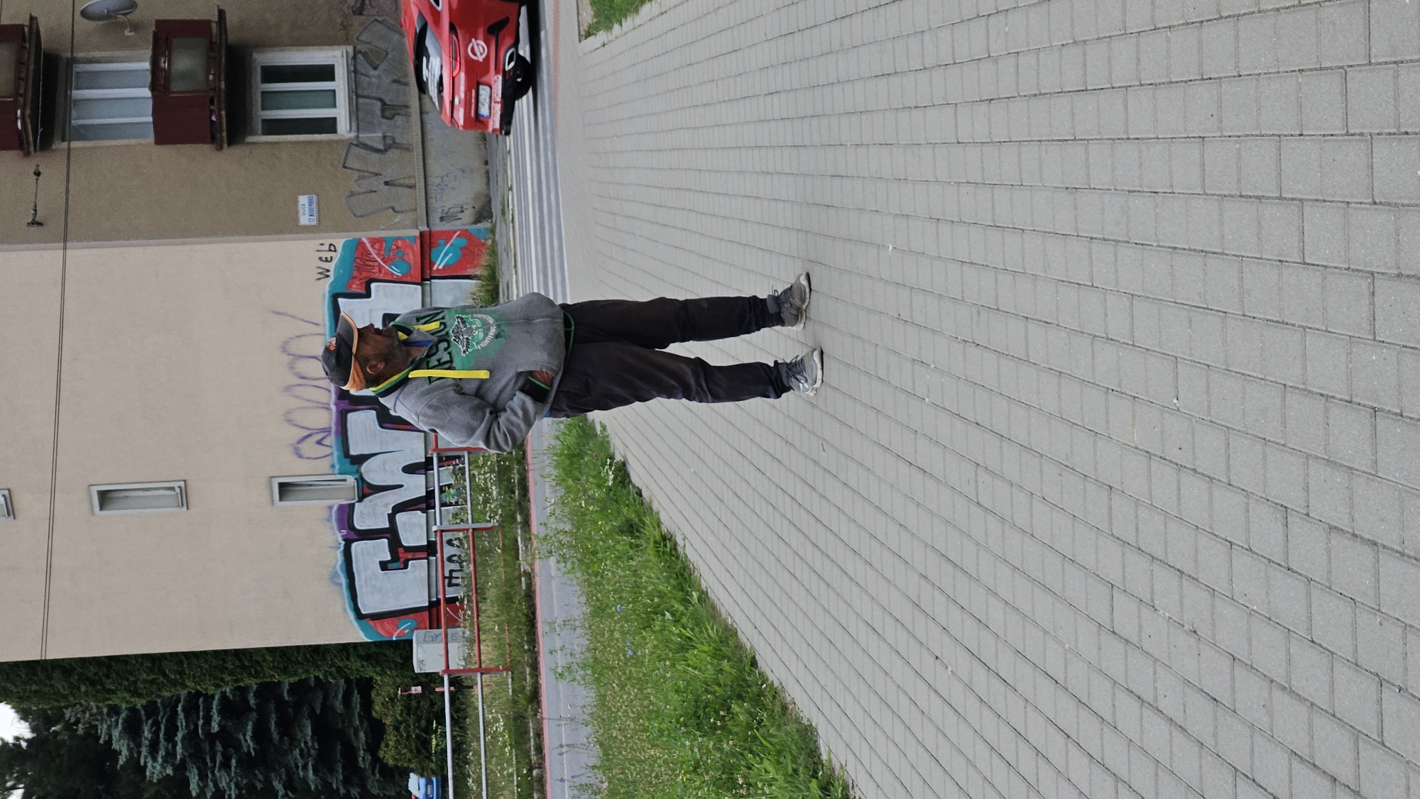 A man wearing a gray hooded sweatshirt with a yellow drawstring, dark pants, a baseball cap, and sneakers stands on a paved sidewalk. He has his hands in his pockets and is looking to his right. Behind him, a building with graffiti is visible, as well as a red car at a crosswalk. This scene is located on Májová Street in Prešov, Slovakia.