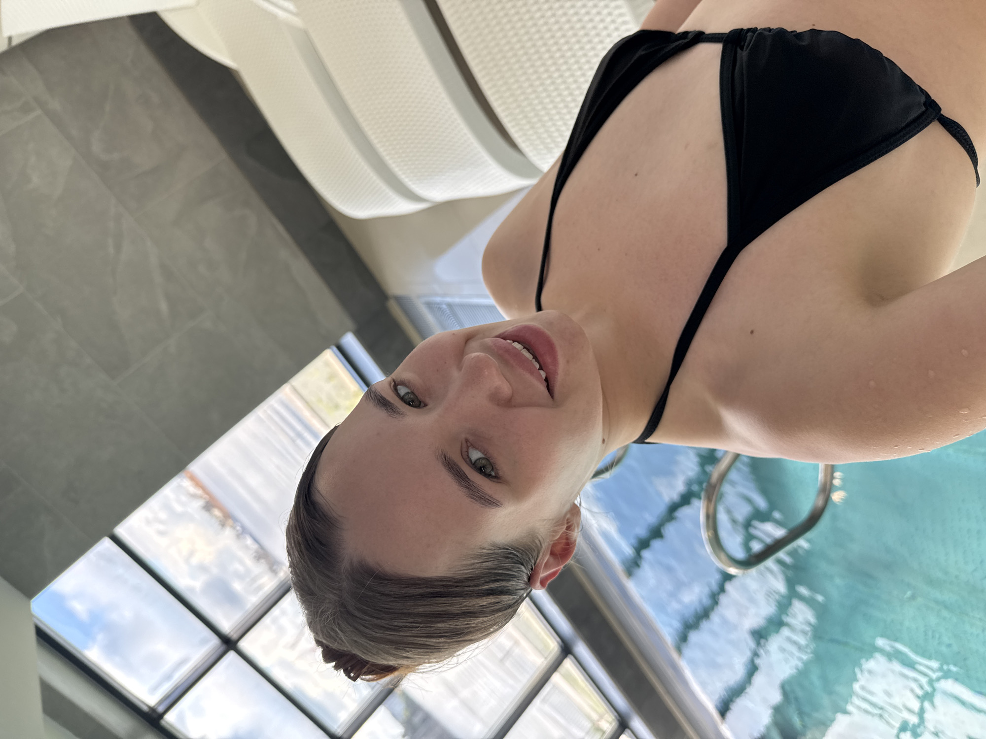 A fair-skinned woman with light brown hair pulled back in a bun is depicted wearing a black bikini top. She is smiling at the camera. The background includes white lounge chairs, gray tile flooring, a window with multiple panes showing a cloudy sky, and what appears to be the edge of a pool with a metal handrail. No specific indication of Chaikovsky, Russia, is visible in the image.