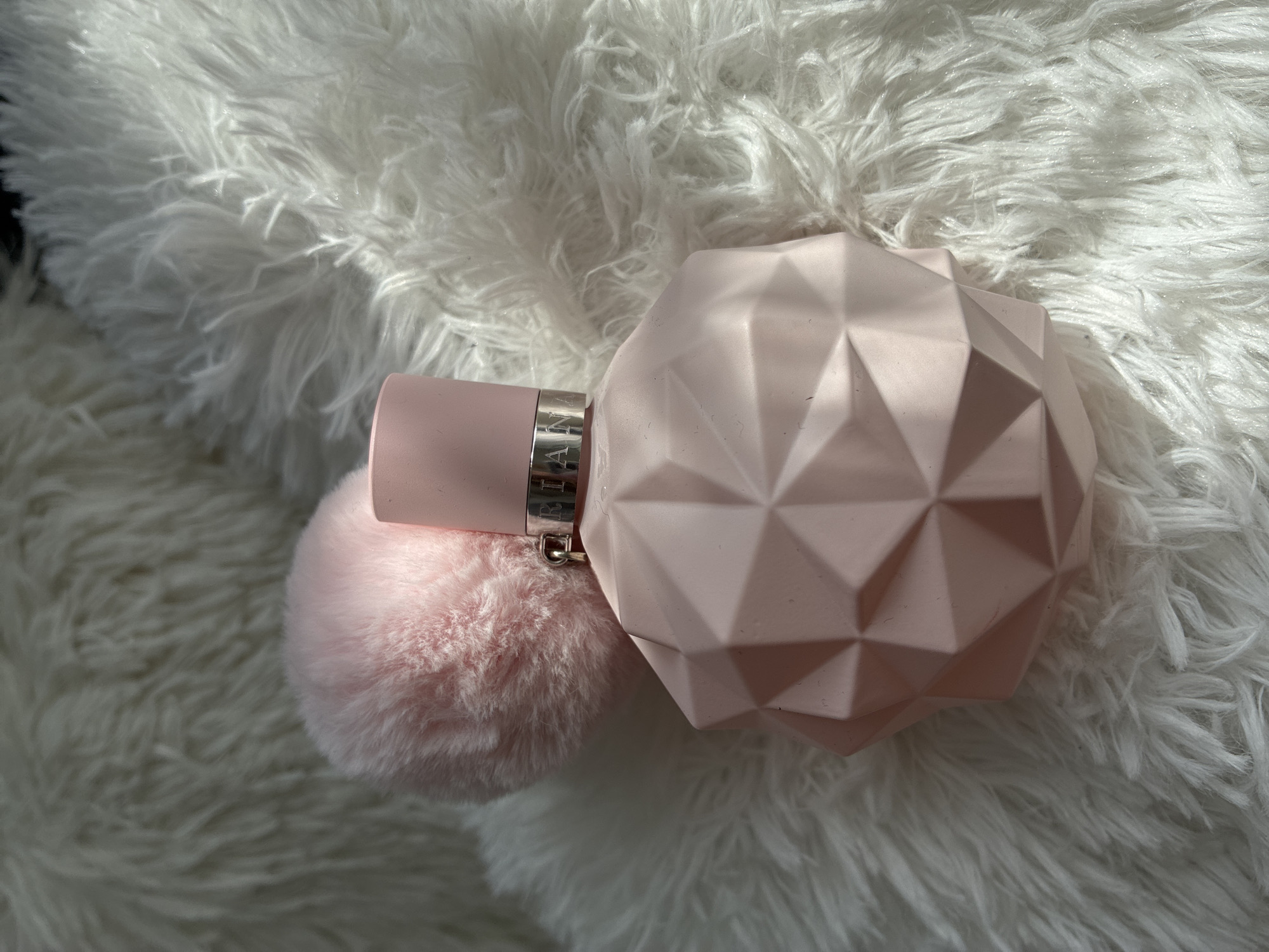 A pink, geometric-shaped perfume bottle is placed on a white, fluffy rug. A light pink pom-pom is positioned behind the perfume bottle. The lighting suggests it is daytime, with sunlight casting shadows on the rug. There is no discernible location in Burgas, Bulgaria.
