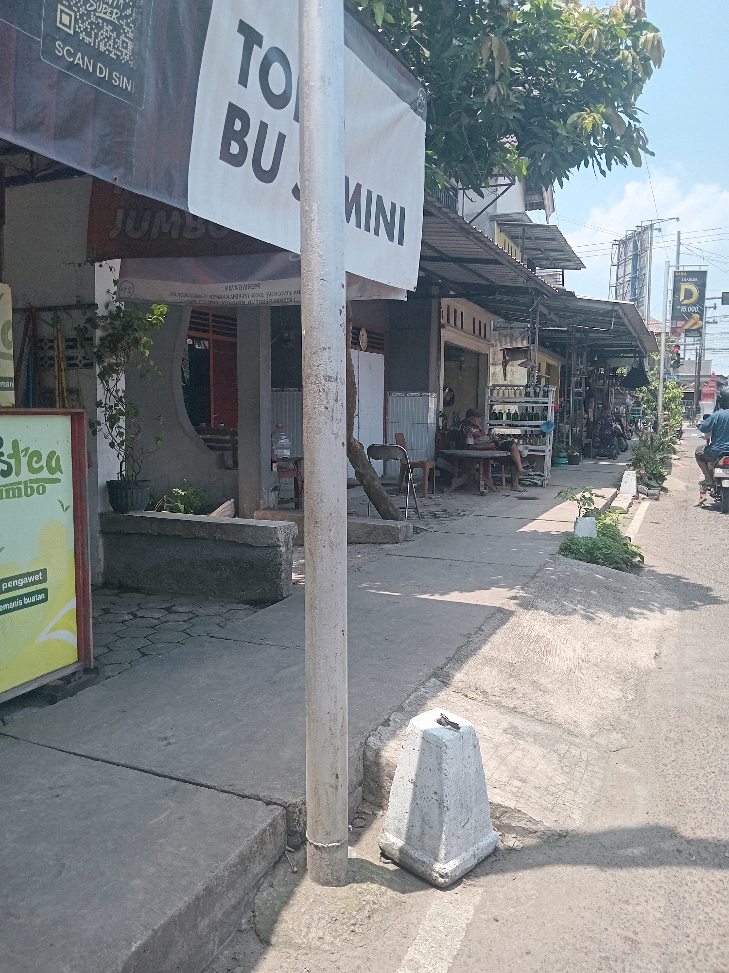 This is a street view image taken in Branggahan, Indonesia. A sidewalk runs along a street with shops and businesses on the right. A large white banner with black lettering "TO BU MINI" is prominently displayed across the top of the frame, partially obscuring the businesses. Below the banner, a sign for "JUMBO" is visible. On the left side of the sidewalk, a green and yellow sign advertises "stca Jumbo".
The sidewalk is made of concrete, with cracks and uneven sections. A white cylindrical pole stands near the foreground on the right. Further down the sidewalk, a concrete barrier is placed on the edge of the street. Several tables and chairs are set up outside one of the shops, where a person is seated. Behind the tables, shelves display rows of bottles. In the distance, a traffic light and a billboard are visible. A person on a motorcycle is riding on the street to the right. The sky is clear with some clouds. Trees and foliage are present in the scene, casting shadows on the sidewalk.