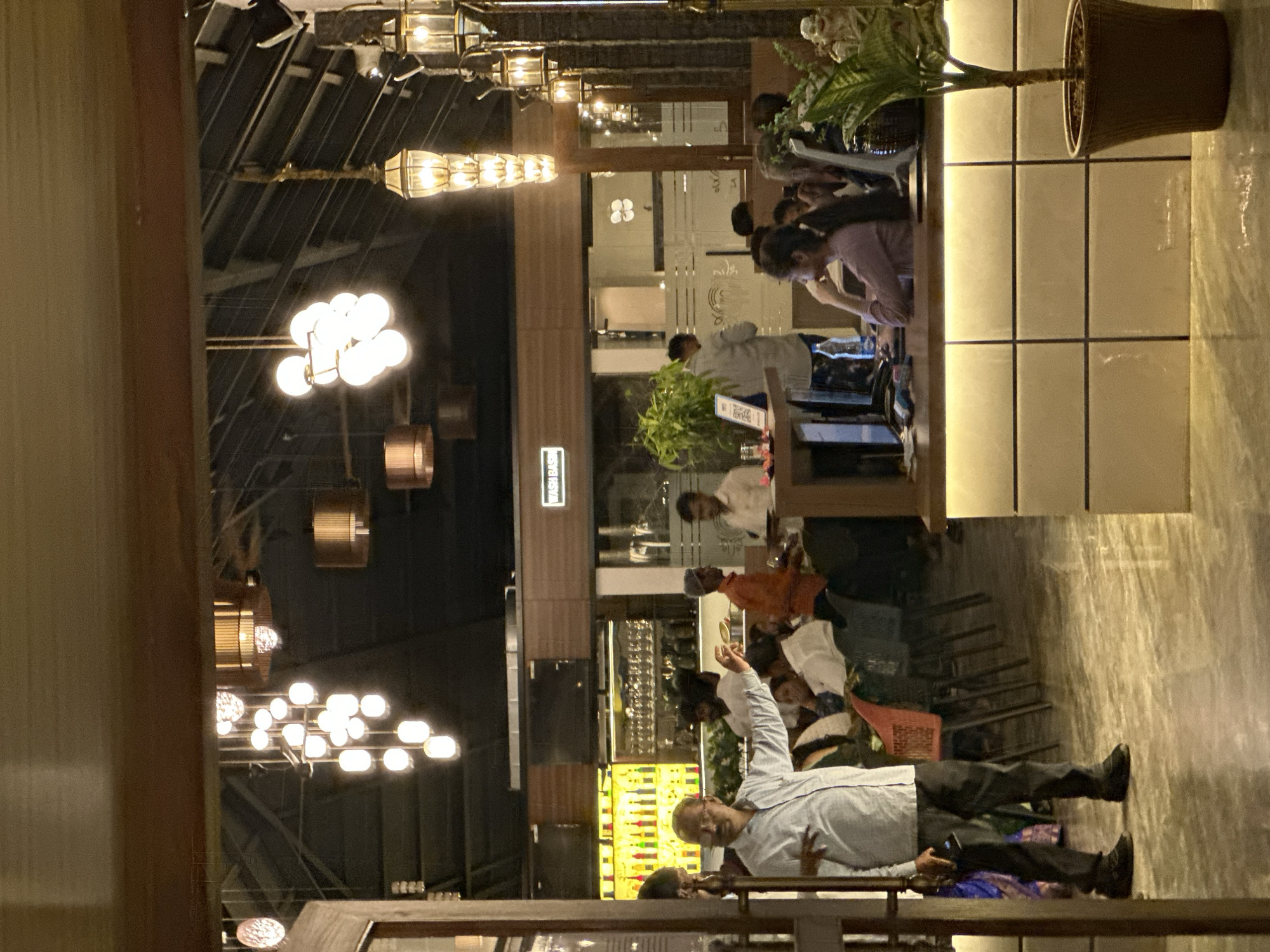 The image captures the interior of what appears to be a restaurant or dining establishment. Several light fixtures are present in the ceiling, including decorative chandeliers. People are seated at tables, suggesting a dining activity. A counter area with a potted plant on the side can be seen. In the foreground, a person is standing with one hand raised. The scene gives the impression of a somewhat crowded restaurant. There is no definitive indication of location within Kalamb, India.