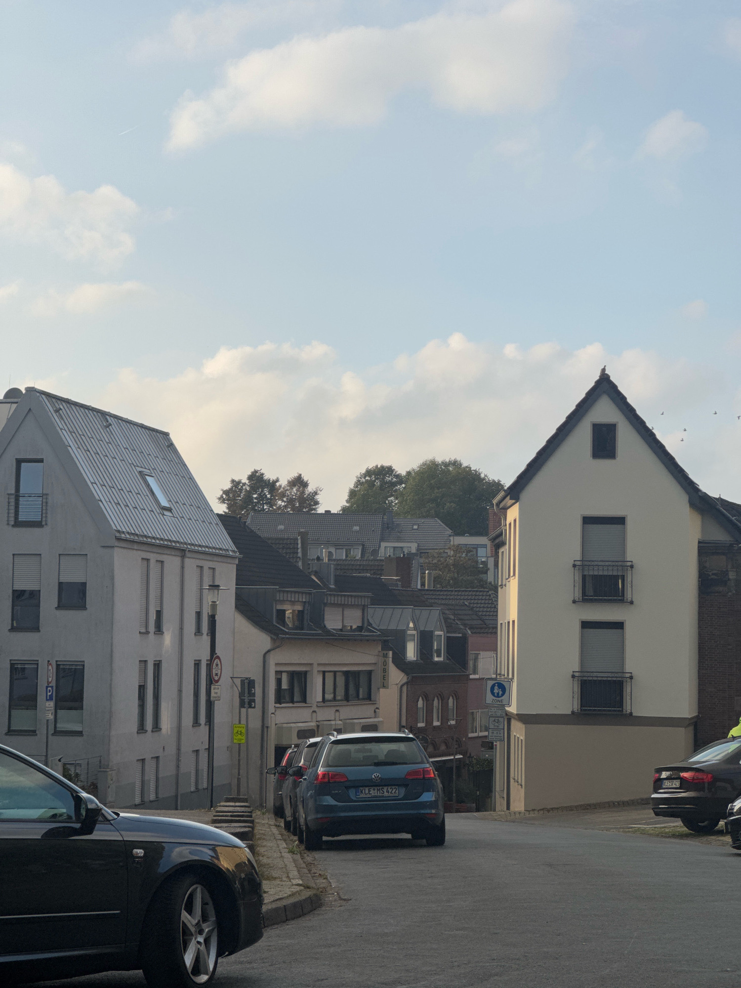 The image depicts a street scene in the city of Cleves, Germany. The perspective is from a slightly elevated position, looking down a gently sloping street lined with multi-story buildings. The sky is a pale blue with scattered white clouds, and a faint contrail is visible.
On the left side of the street is a light-colored, multi-story building with a grey, sloped roof. It features several windows with closed blinds and a small balcony. Adjacent to it, further down the street, are other buildings of varying architectural styles and colors.
On the right side of the street, a prominent building with a cream-colored facade and a dark, sloped roof stands out. This building also has several windows, some with balconies.
Several cars are parked or driving on the street. In the foreground, on the left, a dark-colored car is partially visible, its front end facing away from the camera. Further down the street, a blue hatchback and a black sedan are parked. A pedestrian crossing sign and a sign indicating a bicycle zone are visible on the right side of the street. Trees are visible in the background, suggesting a partly urban, partly suburban setting. The overall impression is a quiet residential or mixed-use street.