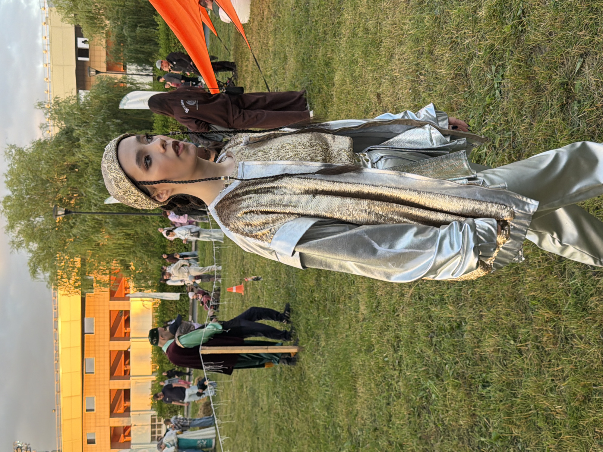 A young woman with dark hair styled in braids stands in a grassy field, looking to her right. She wears a decorative gold headband and a matching intricate top with a shimmering silver jacket over it, adorned with gold accents. Her pants are also silver and shiny. In the background, several people are gathered, some in traditional clothing, and a building with orange accents is visible. The scene appears to be outdoors in a park or event space, with trees and streetlights present. The lighting suggests it is late afternoon or early evening, with a cloudy sky overhead.