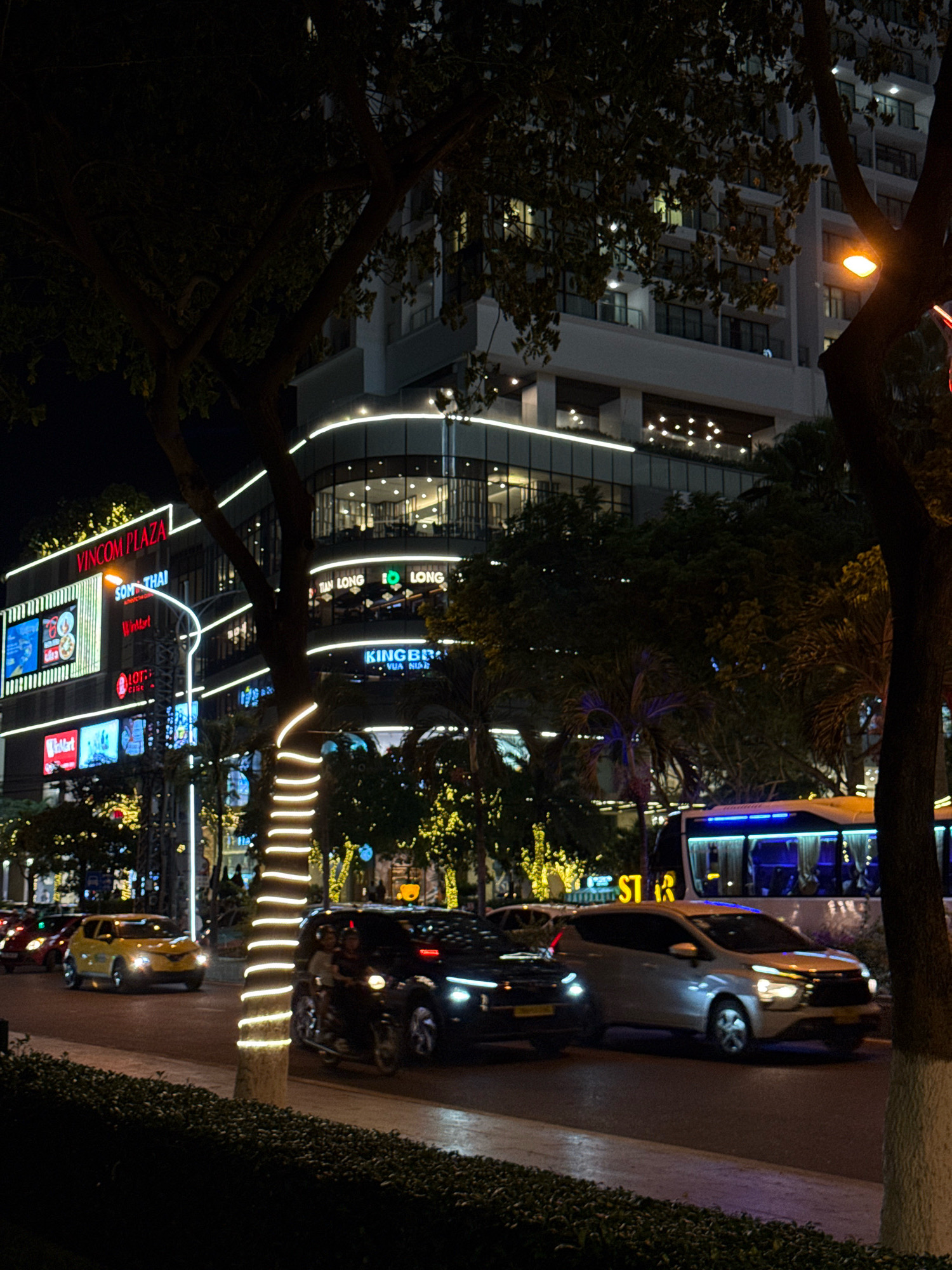 The image depicts a nighttime street scene in Nha Trang, Vietnam, specifically along the Vincom Plaza. A multistory building with illuminated signage and architectural lighting dominates the background. Visible signs include "VINCOM PLAZA," "SON THAI," "LOTTE CINEMA," "Winkart," "TAN LONG," and "KINGBRAND." Several vehicles are present on the road, including a yellow car in the foreground, a black car, a white car, and a motorcycle with two occupants. A large bus with blue interior lighting is parked or moving on the right side of the image. Trees with illuminated trunks and foliage, as well as decorative string lights, line the street, creating a festive atmosphere. The street is paved, and a manicured hedge is visible in the lower foreground. The scene captures the vibrant commercial and street life of the city at night.