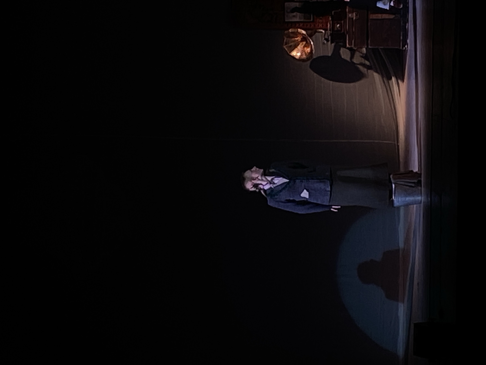 The image shows a theatrical stage scene, presumably in a city like Rusanovo, Russia. A spotlight illuminates a woman lying on the stage, dressed in a dark jacket and skirt. A bucket sits near her. Above her, a complex lighting rig and set decorations cast shadows. The overall environment is dark except for the lit areas highlighting the performer.