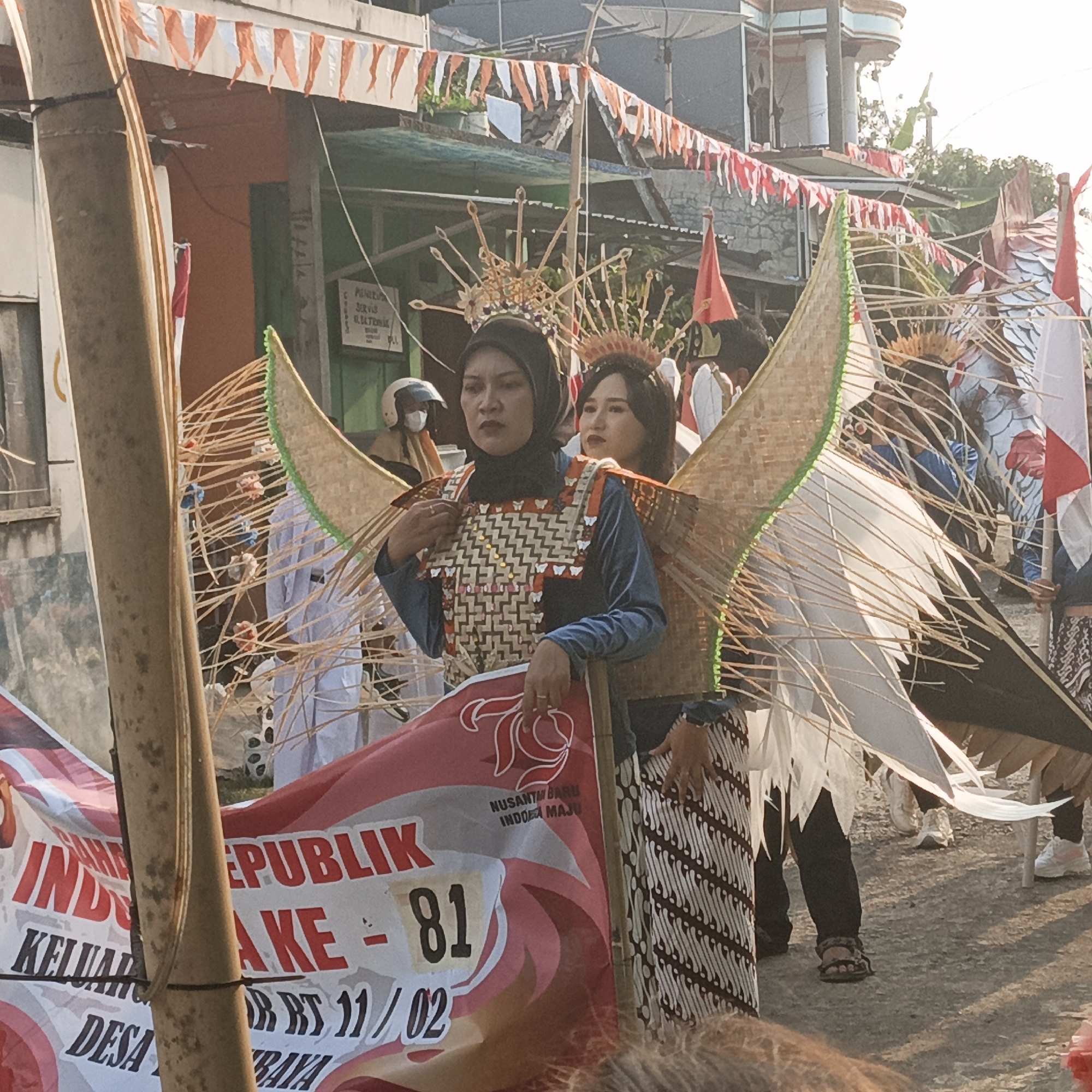 In Tanahbaya, Indonesia, a festive procession is underway, indicated by the red and white bunting strung across the scene. The focal point is two individuals adorned in elaborate costumes. The woman in the foreground wears a black hijab and a chest piece constructed from woven brown and white material, accented with what appear to be small flowers. She has large, wing-like structures made of bamboo and fabric extending from her back. Behind her, another woman in a dark hijab and a similar decorative headpiece is visible, also with wing-like embellishments. They stand near a large red banner with white lettering, parts of which are legible: "REPUBLIK KE - 81", "KELUARGA", and "RT 11 / 02". The banner also displays the text "NUSANTARA BARU INDONESIA MAJU". Other participants in the procession are partially visible, including someone wearing a white outfit and a helmet, and others in patterned batik skirts. The environment appears to be a street, with buildings and trees in the background. The sunlight suggests it is daytime.