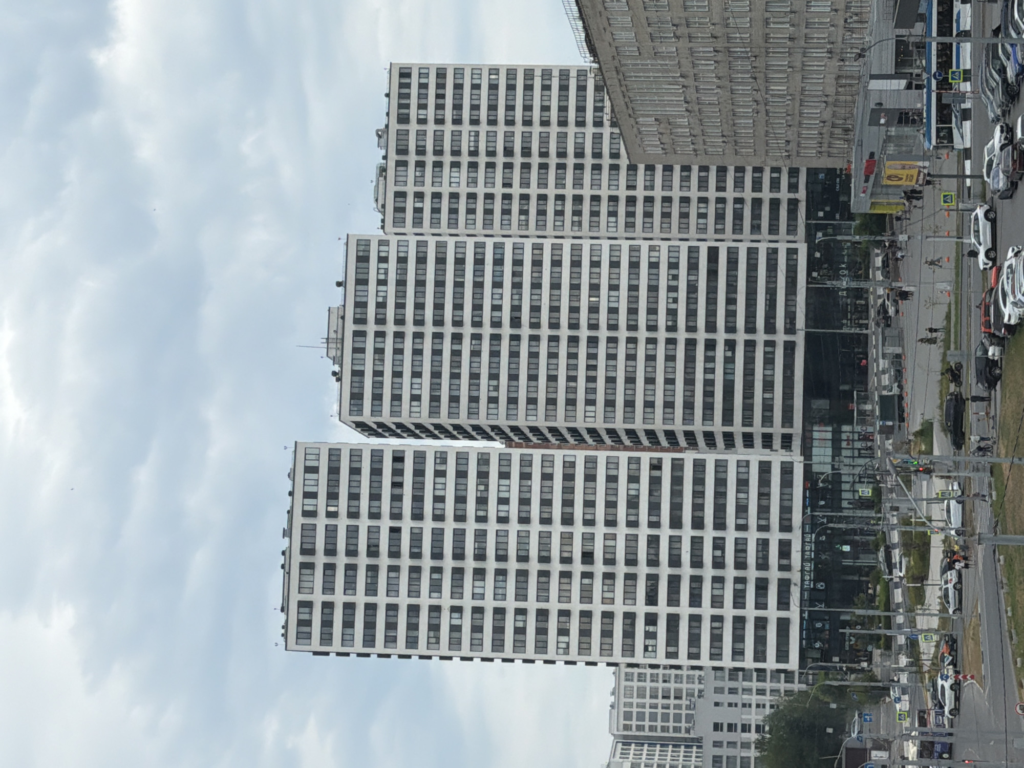 This is an exterior, eye-level, medium-shot photograph of a cityscape in Novosibirsk, Russia, on a cloudy day. The primary focus is on a cluster of modern, tall residential buildings with white facades and numerous dark-paned windows. To the right, a lower, older, grey concrete building with many windows is visible. In the foreground, a street intersection with several cars, pedestrians, and a trolleybus is depicted. Some of the cars are parked, while others appear to be in motion. Pedestrians are walking on sidewalks and crossing the street. The environment includes streetlights, traffic signals, and some greenery. The sky is overcast with scattered clouds. There is no indication of any specific event or action of note.