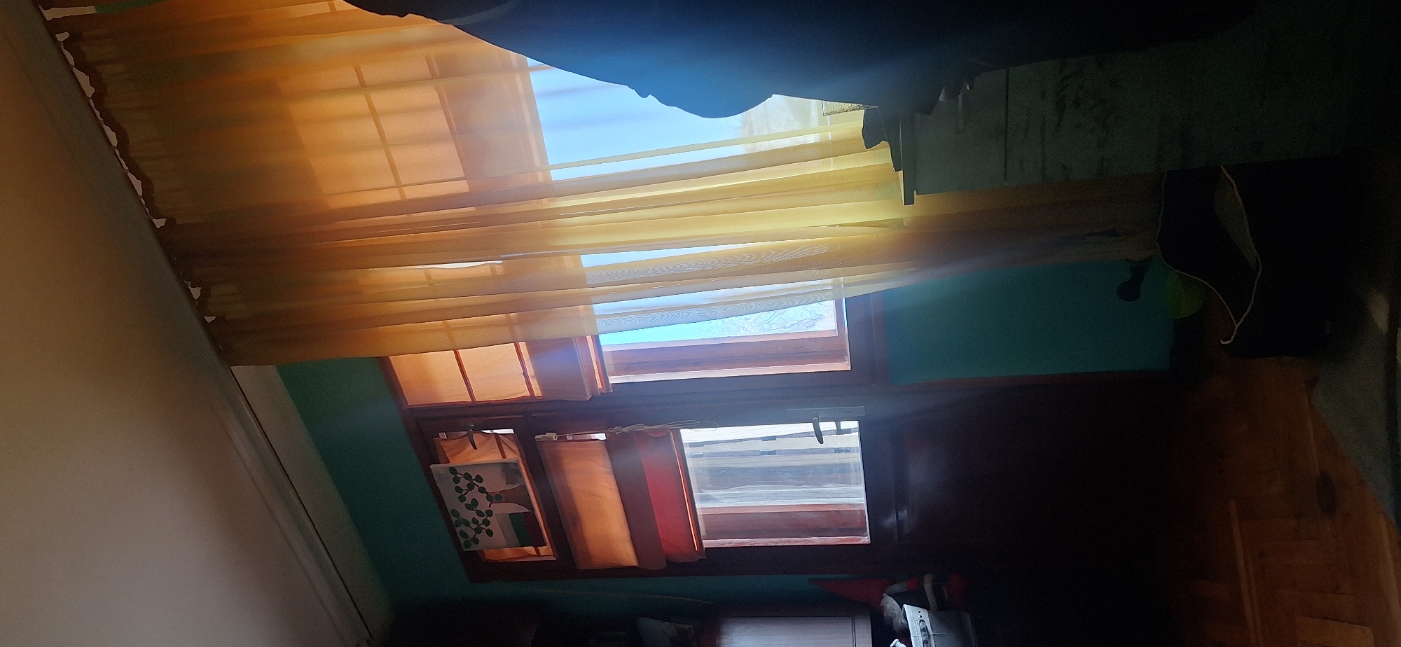 The image depicts an interior scene with two adjacent windows. Both windows are covered by transparent, gold-colored curtains. The upper window reveals a view of a clear, blue sky. The lower window has an additional layer of orange and red curtains. On the left side of the lower window hangs a picture with a foliage pattern. The wall is painted with a shade of teal. The furniture and floor are primarily brown wood. A black bag hangs on the wall. The image does not include specific landmarks that can identify the location as Stara Zagora, Bulgaria. No people or actions are visible in the image.