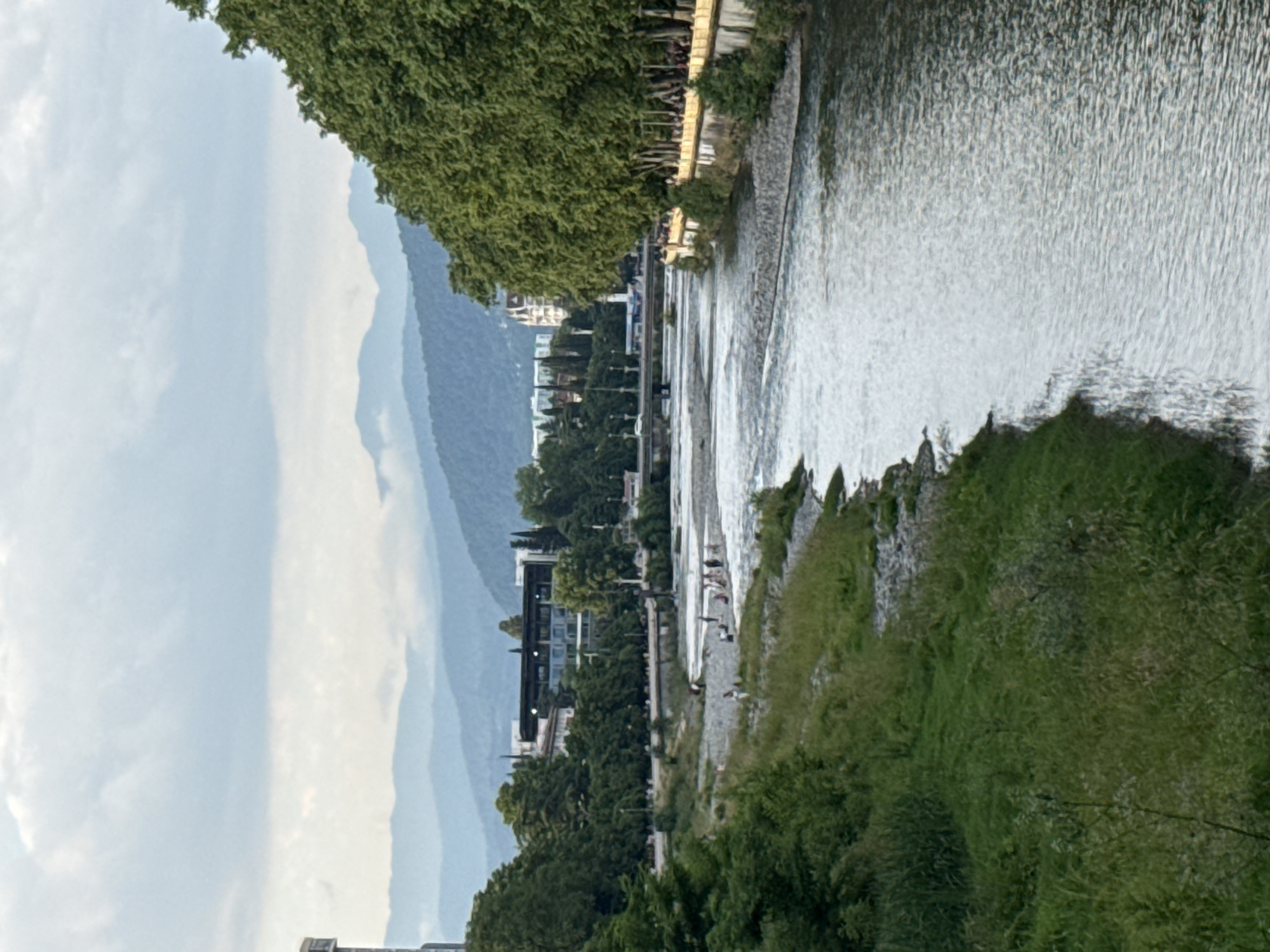 The media file depicts a picturesque scene along a river, likely in Sochi, Russia. In the foreground, the flowing river water creates a textured surface, reflecting the light. The riverbank is lined with lush green vegetation, including trees and grasses, suggesting a natural or park-like setting.
Further into the scene, a paved walkway runs alongside the river, and several people can be seen strolling or standing on it. Beyond the walkway, there are modern buildings with glass facades, interspersed with more trees. The architecture suggests a developed area adjacent to the natural landscape.
In the background, a series of mountains rise, their slopes covered in a darker green foliage. The mountains fade into the distance, creating a sense of depth. The sky above is partly cloudy, with patches of light blue and white clouds, indicating daytime with moderate weather.
The overall atmosphere is serene and offers a view of a well-integrated urban and natural environment. The presence of people suggests a recreational or public space. The time of day appears to be daylight, possibly late afternoon or early evening given the soft light. There is no visible text in the image.