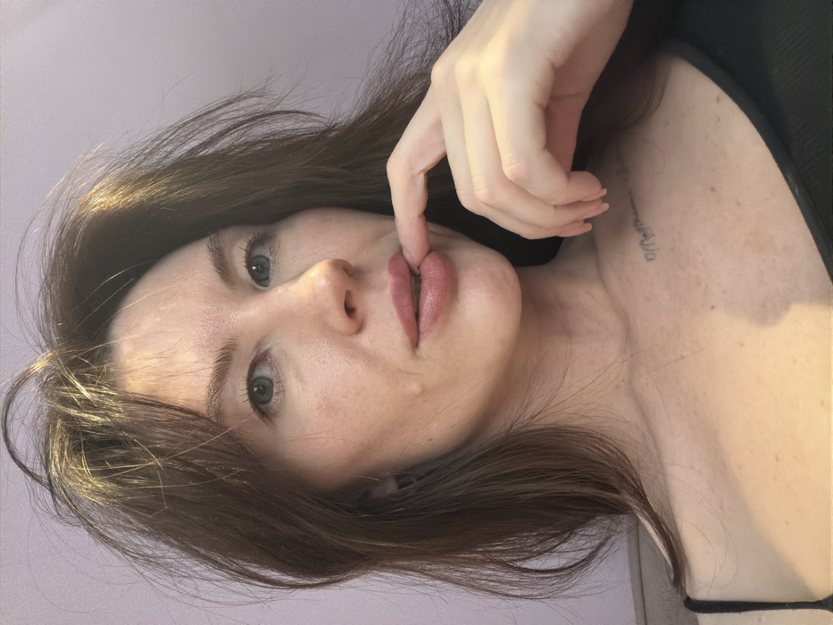 A single adult female is depicted in a close-up, chest-up portrait. She has fair skin, light-colored eyes, and long, disheveled brown hair that frames her face and shoulders. Her right index finger is positioned on her lower lip. She is wearing a dark, possibly black, spaghetti strap top. A faint, dark-ink tattoo is visible on her upper right chest, featuring script-like text. The background consists of a solid light lavender-colored wall, with a dark, indistinct object partially visible on the lower left, possibly furniture. There are no identifiable features within the image to determine a specific location in Perm, Russia, or any other city.