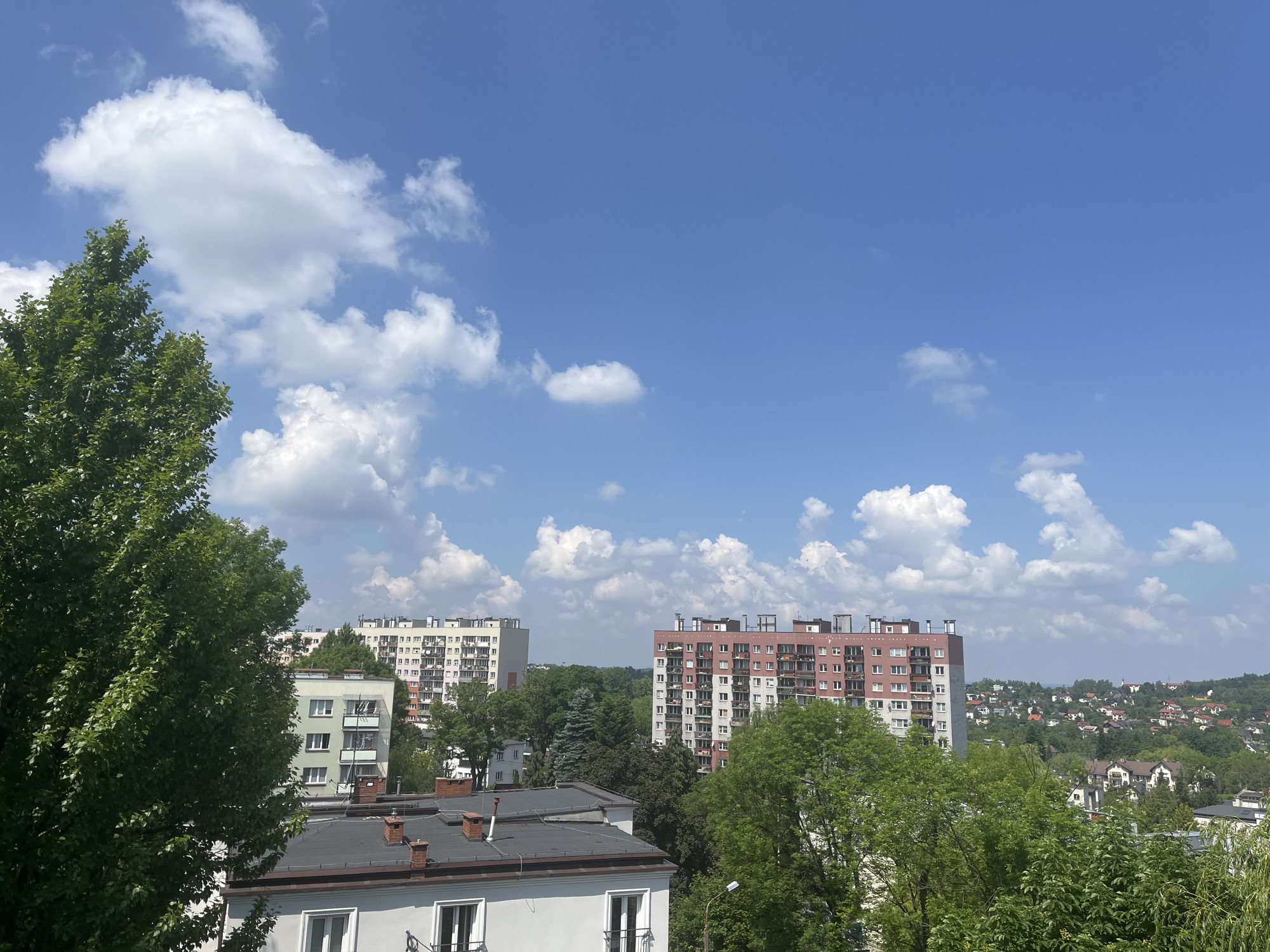 This image captures a bright, sunny daytime scene of a residential area, showcasing a blend of urban architecture and natural greenery under a clear blue sky.
The sky, occupying the upper half of the frame, is a vivid azure, dotted with numerous fluffy, white cumulus clouds. The bright sunlight suggests fair weather, casting gentle illumination across the landscape.
In the foreground, the dark, pitched roofs and chimneys of several lower-rise buildings are visible, indicating residential structures. One building's light-colored facade and windows are partially seen. A large, deciduous tree with dense green foliage dominates the left side of the foreground, its branches extending upwards and across.
Further back, the scene unfolds into a more expansive urban landscape. Two prominent multi-story apartment buildings stand out. The one on the left is a light-colored, tall block with many balconies. To its right, another large apartment building features a distinctive design with a light base and an upper section painted in a rust-red or terracotta color, also adorned with numerous balconies.
Between and around these buildings, substantial amounts of green trees and bushes are present, indicating a well-vegetated environment. In the distant background, rolling hills are covered with a sprawling residential area consisting of many smaller houses with visible red roofs, extending towards the horizon.
No specific human activity or event is taking place, and no discernible text is visible anywhere in the image. The overall impression is one of a serene, populated area enjoying a pleasant day.