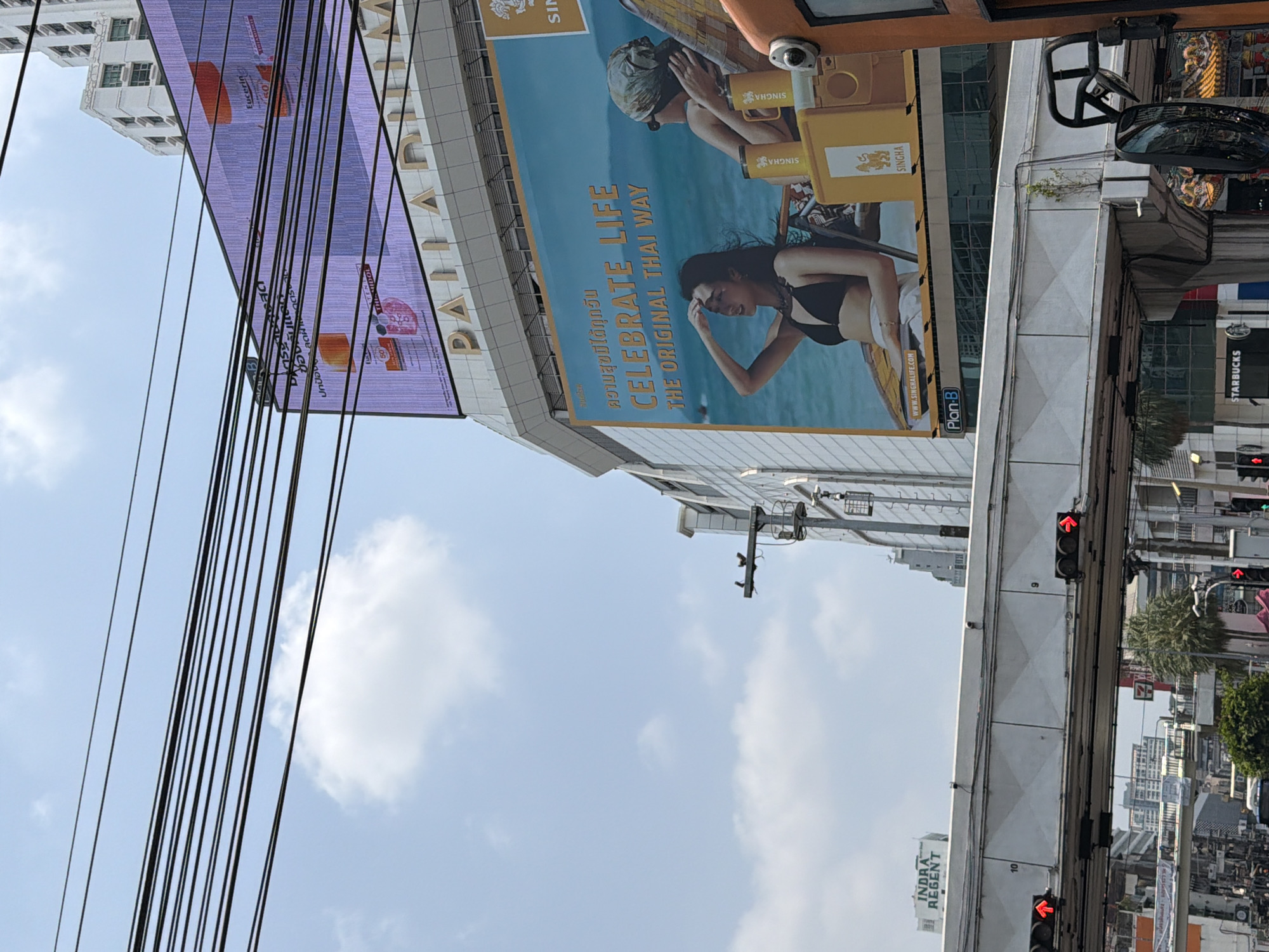 The image captures a street scene in Bangkok, Thailand. A large billboard promoting "Singha" beer is prominently displayed, featuring a woman on a boat, next to another advertisement. A digital billboard with advertisements for Eucerin is also visible. Utility lines crisscross the sky. Lower down, an elevated train line runs along the street, with traffic lights visible. The "Indra Regent" building can also be spotted. In the background, other buildings suggest a densely populated urban environment. A Starbucks sign can also be seen.