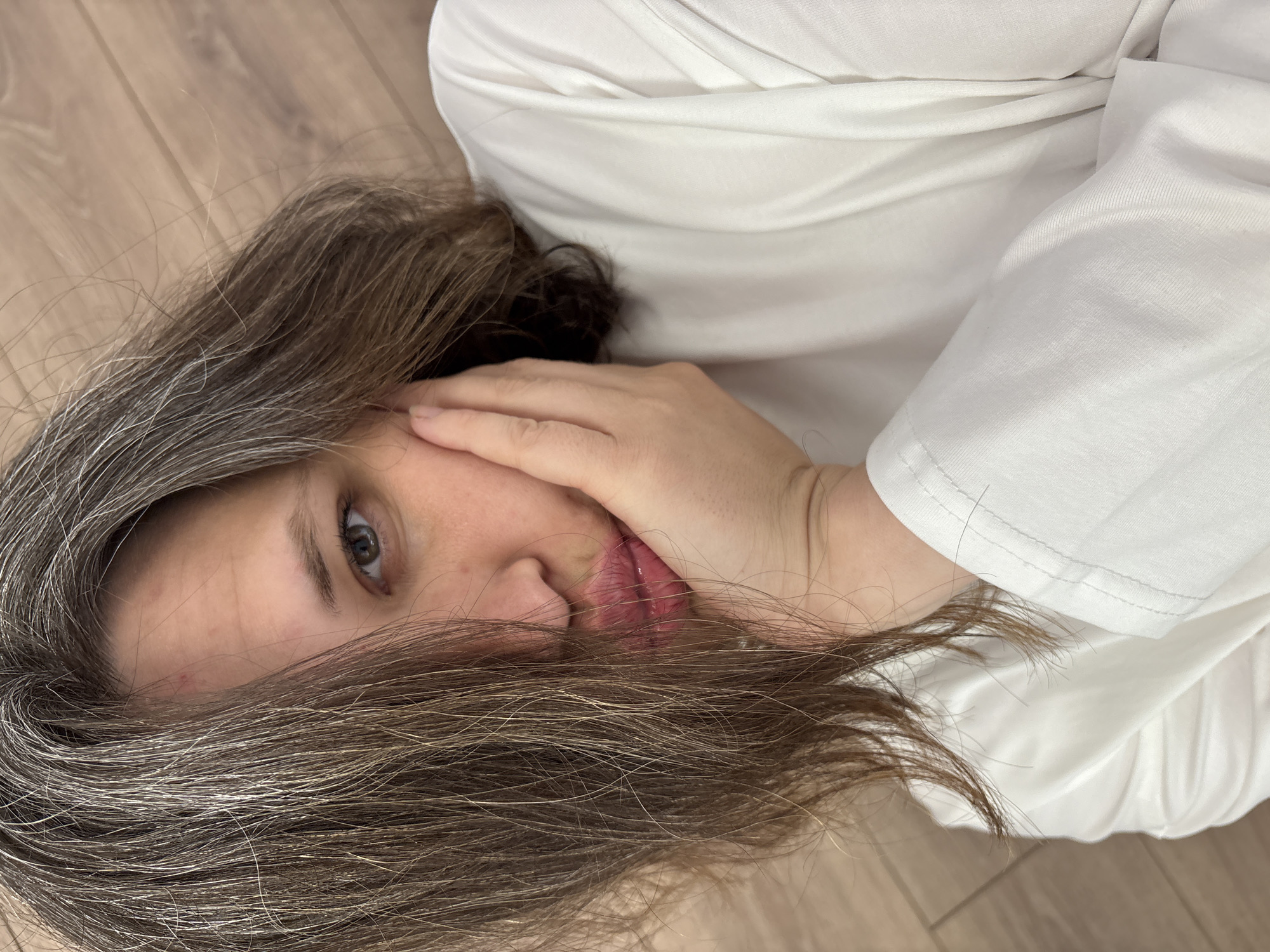 A woman with brown and grey hair is lying on a wooden floor. She is wearing a white shirt. Her right hand is covering part of her face. The environment appears to be indoors. The location is not identifiable from the image.