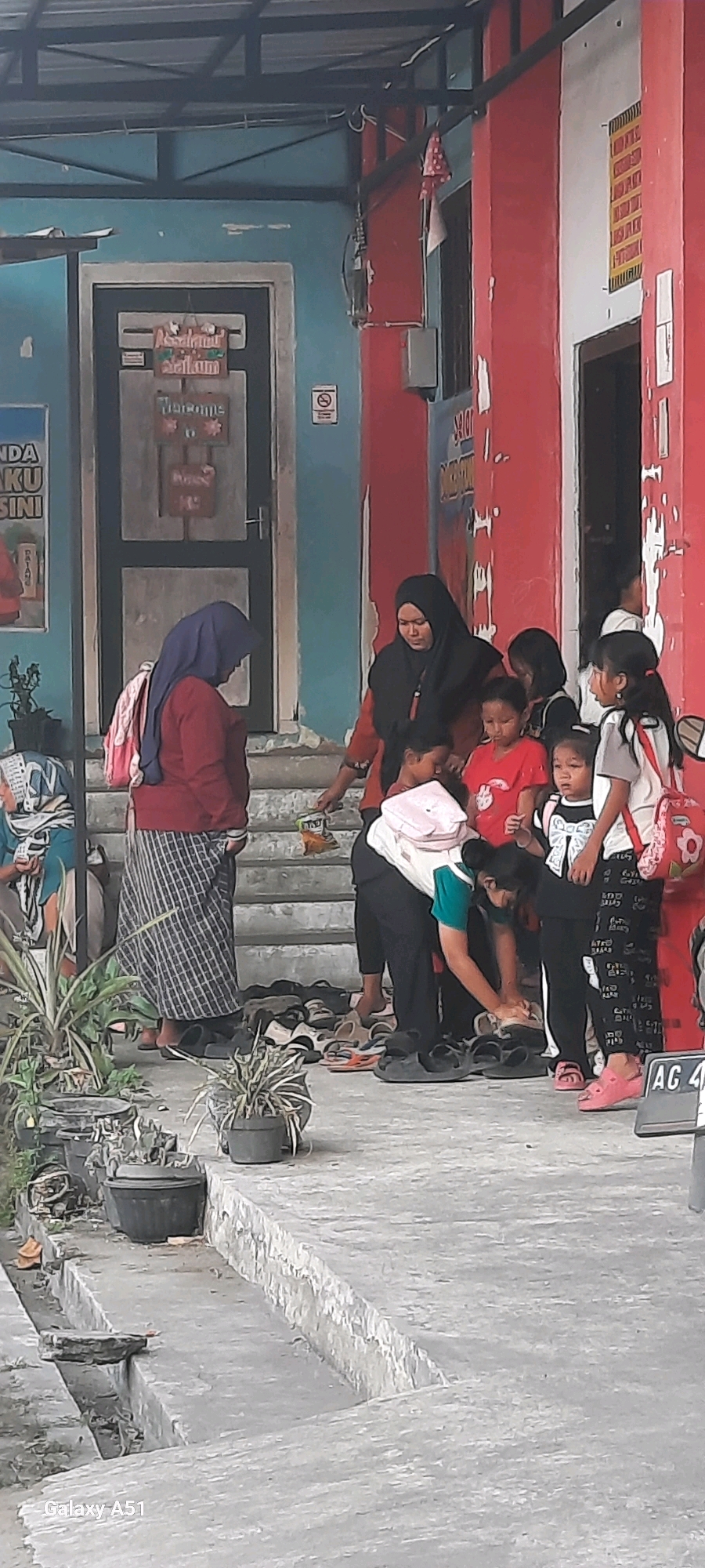 The image depicts an outdoor scene in front of a building with a red and blue facade, potentially located in Selopuro, Indonesia. Several individuals are present, including adults and children, gathered on a concrete step area. Some individuals appear to be adjusting or putting on footwear, with a collection of sandals and shoes visible on the ground. A woman wearing a black hijab and an orange top is interacting with a group of children. Another woman, wearing a blue hijab and a red top, stands to the side with a backpack. The environment includes potted plants and a sign above a doorway that reads "Assalamualikum" and "Welcome." The concrete ground is uneven, and a drainage channel is visible in the foreground.