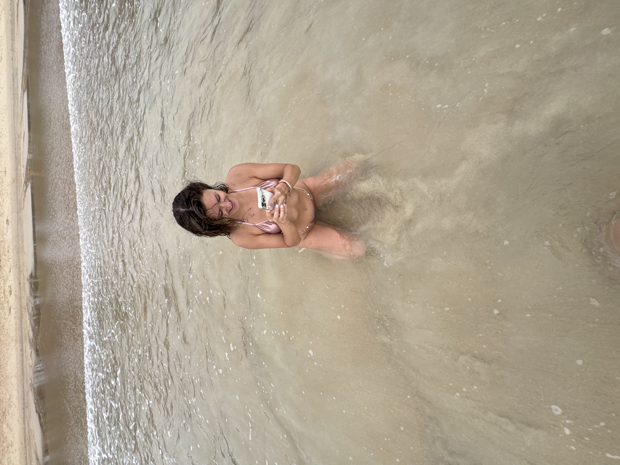A woman is kneeling in shallow water at a beach, looking at her phone. She is wearing a pink bikini. The water is clear with a sandy bottom. The waves are gentle and foamy. The background shows the sandy shore of a beach. This location is likely in Choeng Thale, Thailand, known for its beautiful beaches.
