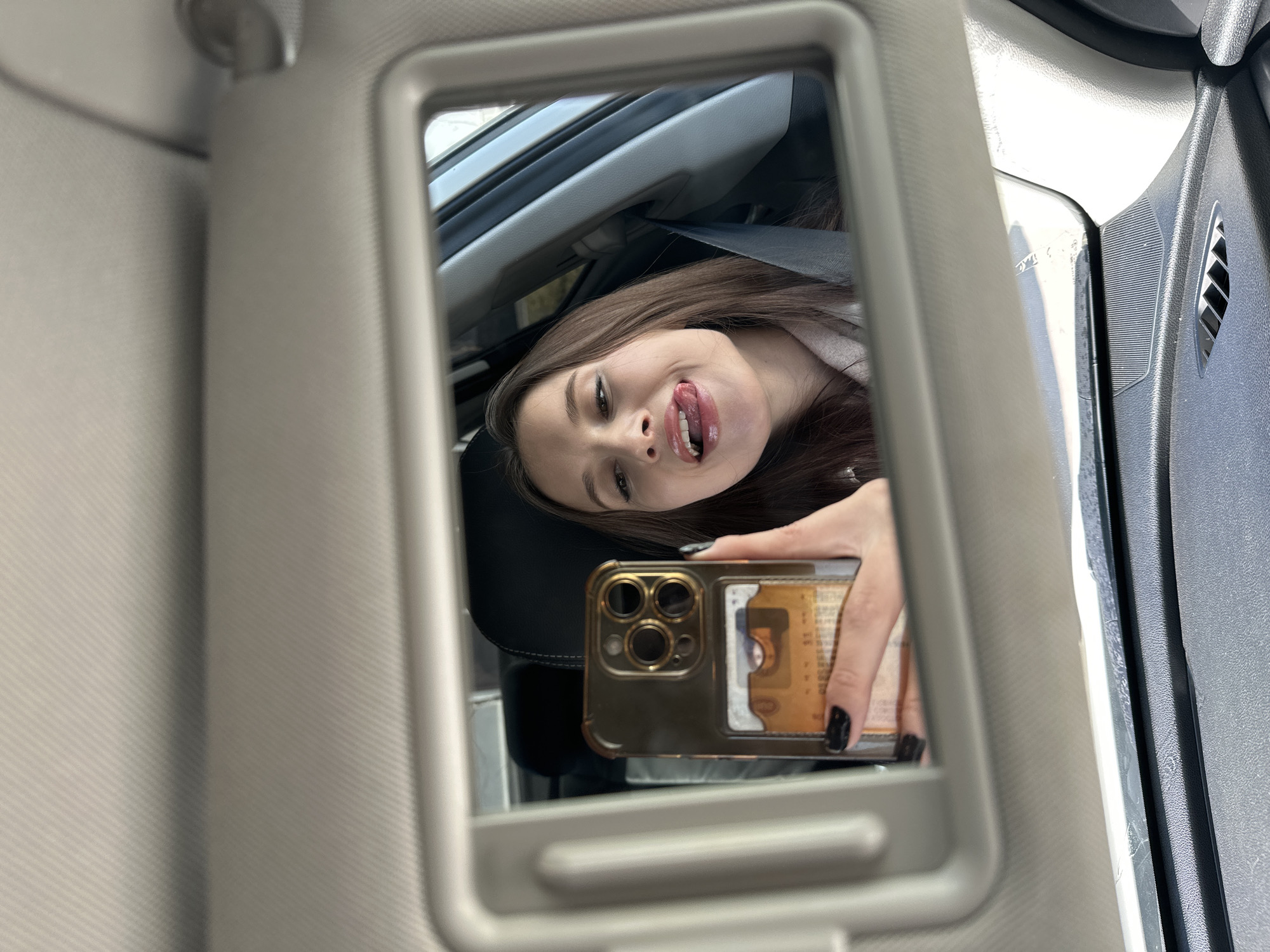 A young woman is captured in a reflection, taking a mirror selfie inside a car in Yekaterinburg, Russia. The image is framed by the vanity mirror embedded in the car's sun visor.
The central subject is the reflection of the woman, who appears to have long, dark brown hair. She is looking directly into the phone held in her hand, which is also reflected in the mirror. Her expression is playful, with her tongue slightly extended out of her mouth. She has dark eyes and is wearing dark nail polish on her visible fingers.
She is holding a gold-colored smartphone, likely an iPhone Pro model due to its distinctive multi-lens camera system. A card holder accessory is attached to the back of the phone, and a card, partially visible, clearly shows the text "Bank of America."
The setting is the interior of a car, seen through the reflection and the surrounding elements. The sun visor is beige or light grey, matching the headliner fabric visible on the left. Parts of a dark car seat, a seatbelt, and elements of the car's dashboard, including a speaker grill and air vent, are discernible around the mirror's frame. Natural daylight suggests it is daytime, casting light into the car's interior. No specific weather conditions are apparent.