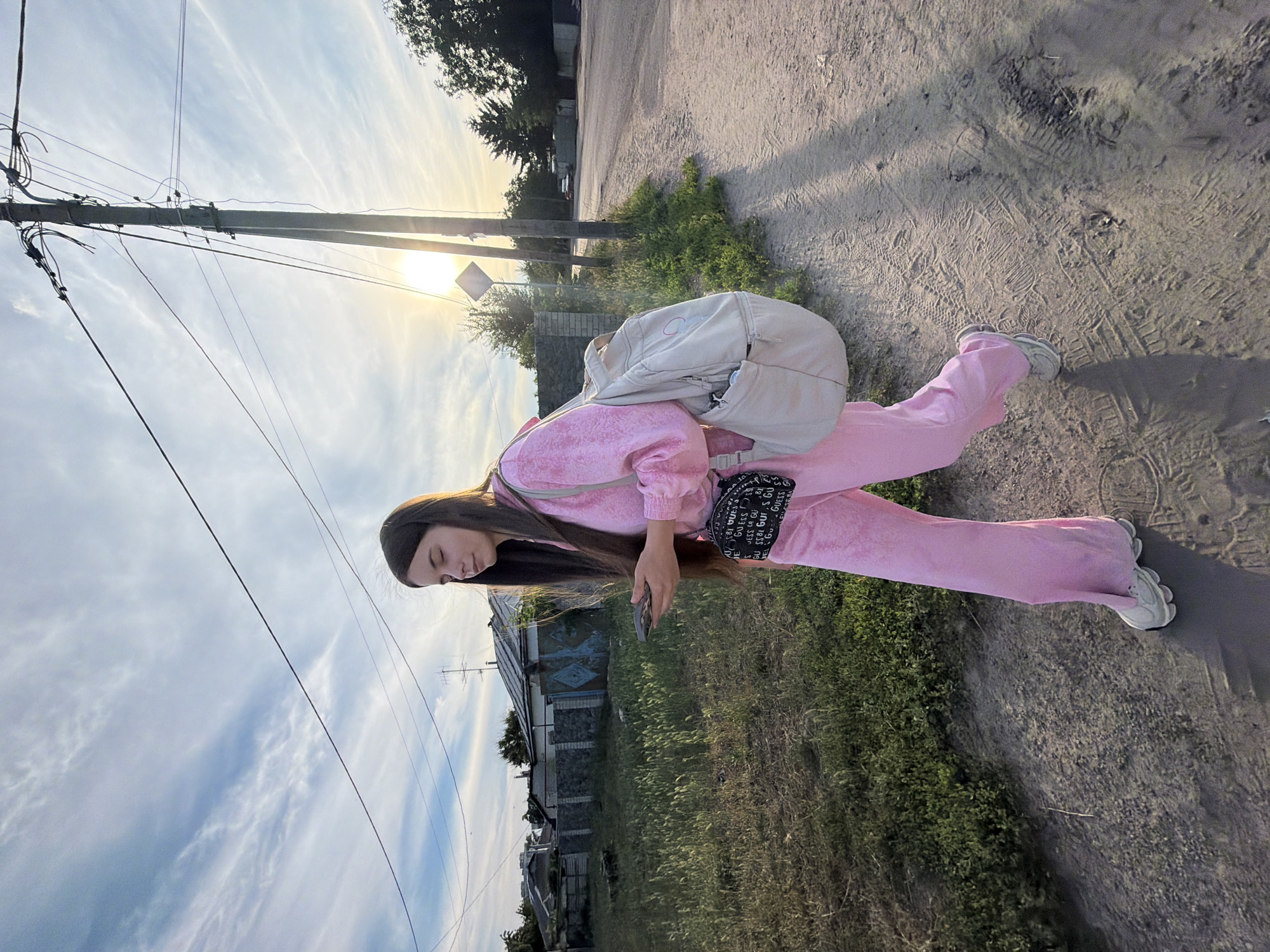 A young woman stands in a dirt path on a sunny day, with a light blue sky and wispy clouds above. She is wearing a pink tracksuit and white sneakers, and has a light-colored backpack slung over her left shoulder, and a small black bag with the word "GUESS" printed repeatedly in white on her right hip. She is looking down at her phone, which she holds in her right hand. In the background, there are houses, trees, and power lines. The sun is setting, casting long shadows on the ground. The scene appears to be in a rural or suburban area, likely in Dnipro, Ukraine. The woman's posture suggests she might be taking a photo of herself or is otherwise engrossed in her phone.