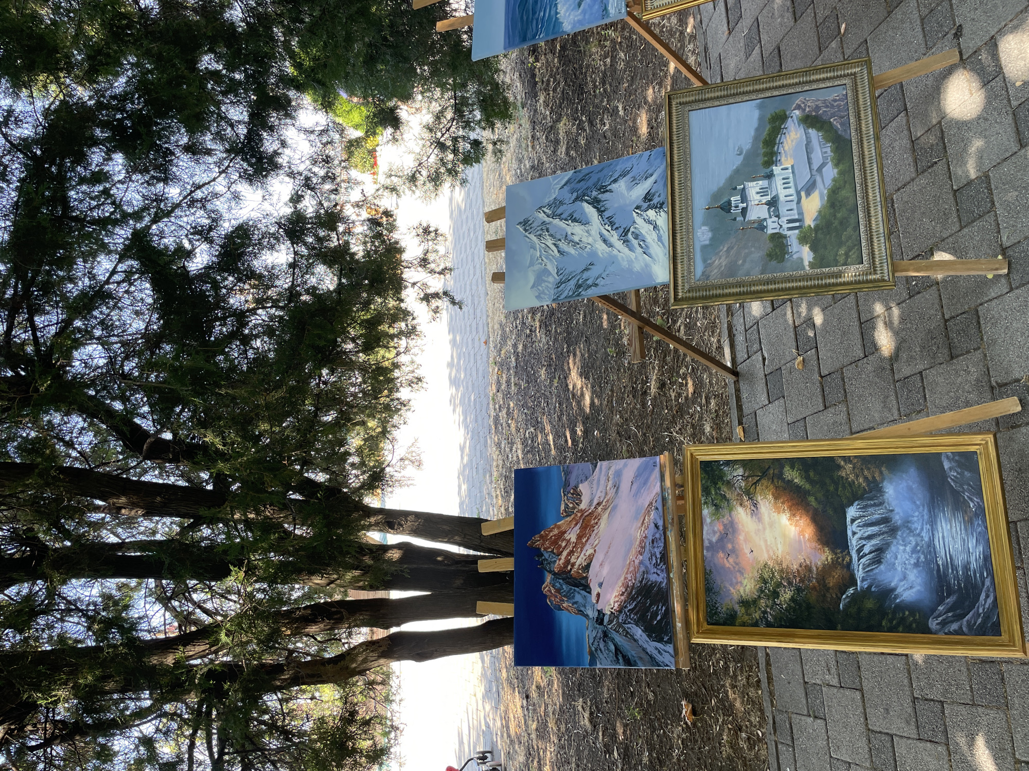 The image shows an outdoor art exhibition featuring several landscape paintings displayed on easels under the shade of trees. The paintings depict various natural scenes, including mountains, a waterfall, and a body of water with waves. In the foreground, there are at least four paintings. One is a framed oil painting of a waterfall with dramatic lighting and birds in the sky. Another framed painting depicts a white domed church situated on a cliff overlooking the sea. Two unframed paintings on easels behind these show a snow-capped mountain range and a dynamic seascape. The ground is paved with grey rectangular tiles, and dry leaves are scattered on the dirt area surrounding the exhibition. The background suggests a sunny day with dappled light filtering through the tree branches. A person pushing a stroller is partially visible in the far left background. The trees appear to be conifers, providing a natural canopy for the display. The scene suggests a public space, possibly a park or promenade, where local artists are showcasing their work. The overall atmosphere is serene and artistic.