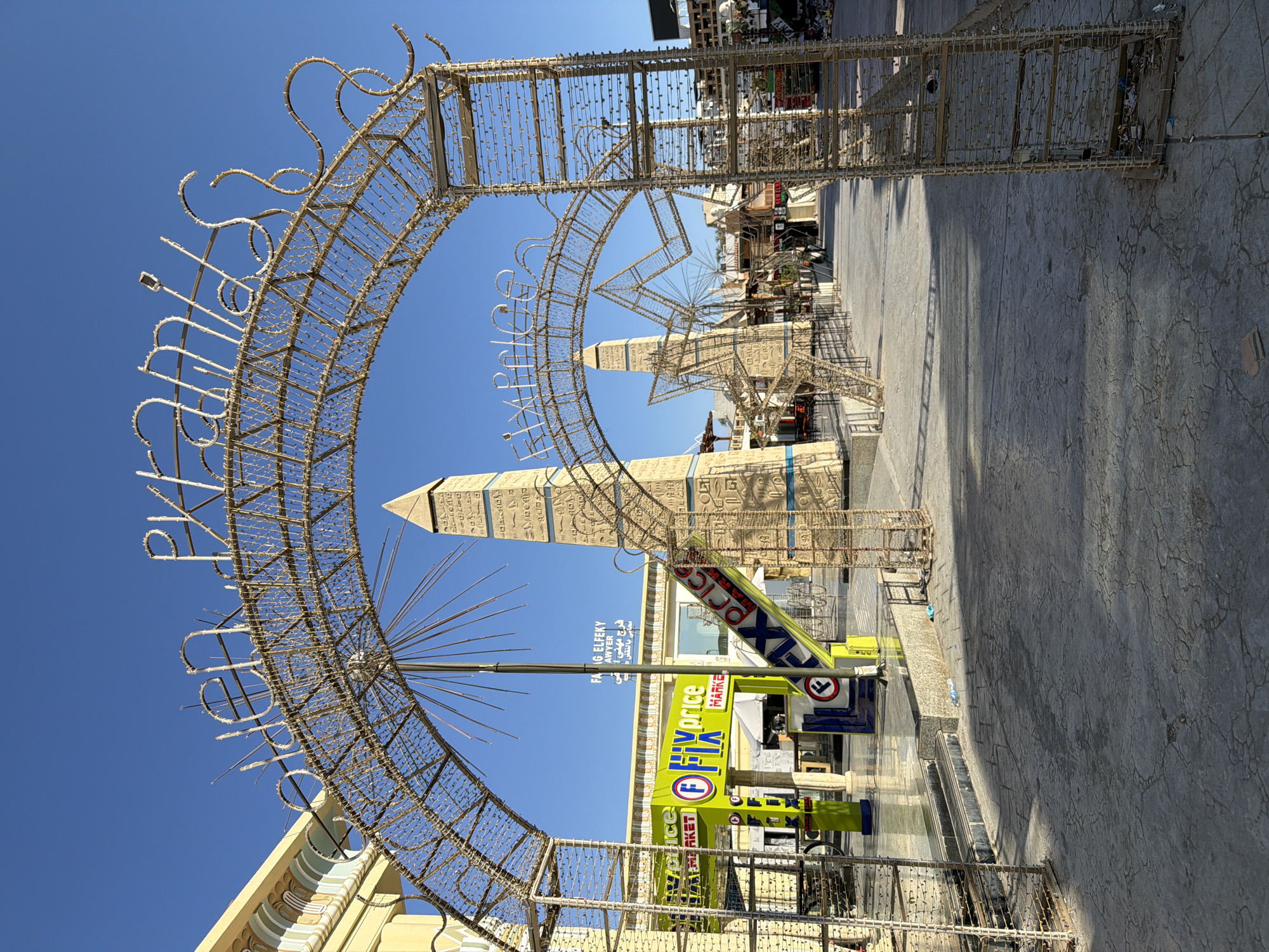 The image captures an outdoor scene in Sharm El Sheikh, Egypt, featuring a decorative arched entryway adorned with lights, spelling out "GOLDEN PYRAMIDS." The entryway is constructed from a metallic lattice framework. In the background, two obelisk-shaped structures, reminiscent of Egyptian monuments, are visible. One obelisk, closer to the foreground, is elaborately decorated with hieroglyph-like carvings and blue accents. To the left, a yellow building with architectural details suggesting an Egyptian theme can be seen. A bright green sign for "FIX PRICE MARKET" is prominently displayed, with a circular red and white logo. The ground is paved with textured concrete. Sunlight casts distinct shadows across the scene. No people are clearly visible in the frame.