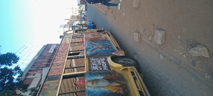 A vibrant yellow matatu, a Kenyan minibus, is parked on the street, facing left. It's adorned with religious imagery and phrases, notably "JESUS MY ONLY HOPE." The surrounding environment includes buildings, a pedestrian walking in the background, and a rough, stone-filled roadside. The building is partially visible on the left with the word "RE" visible. The sky is partially visible.