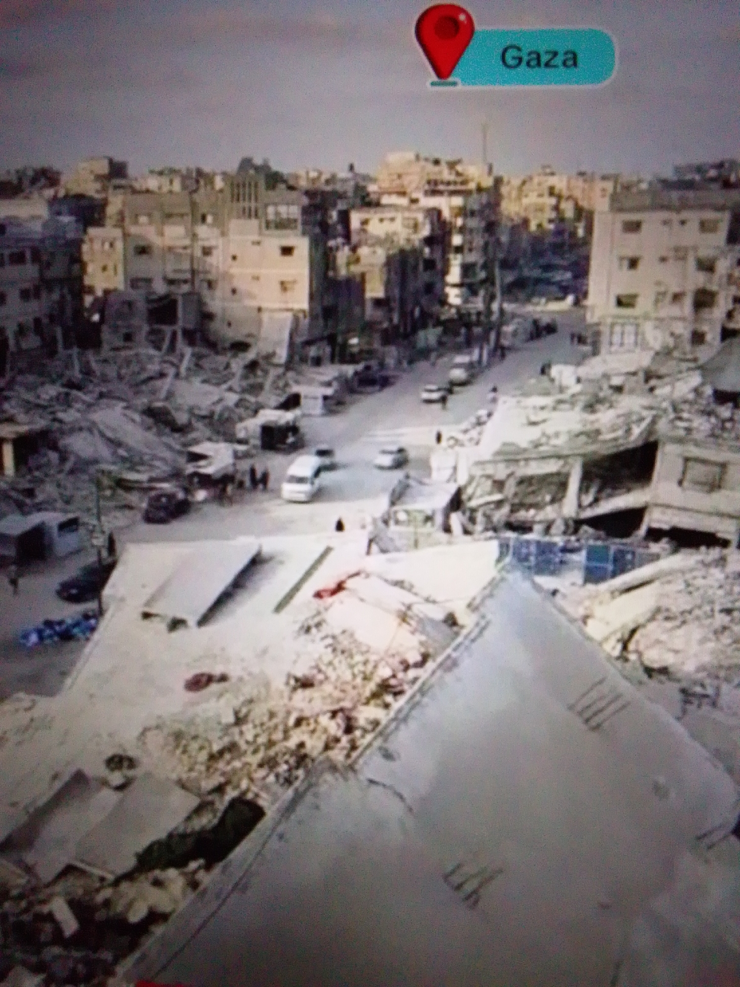 The image depicts widespread destruction in an urban environment. Buildings are heavily damaged, with rubble and debris covering the ground. Several vehicles, including cars and vans, are visible on the streets amidst the devastation. Some individuals are present, appearing to be walking or standing near the damaged structures. A prominent graphic overlay indicates the location as "Gaza." The overall scene suggests a catastrophic event, likely resulting from conflict or a natural disaster. There is no visual information within the image that connects it to the city of Aznakayevo, Russia.