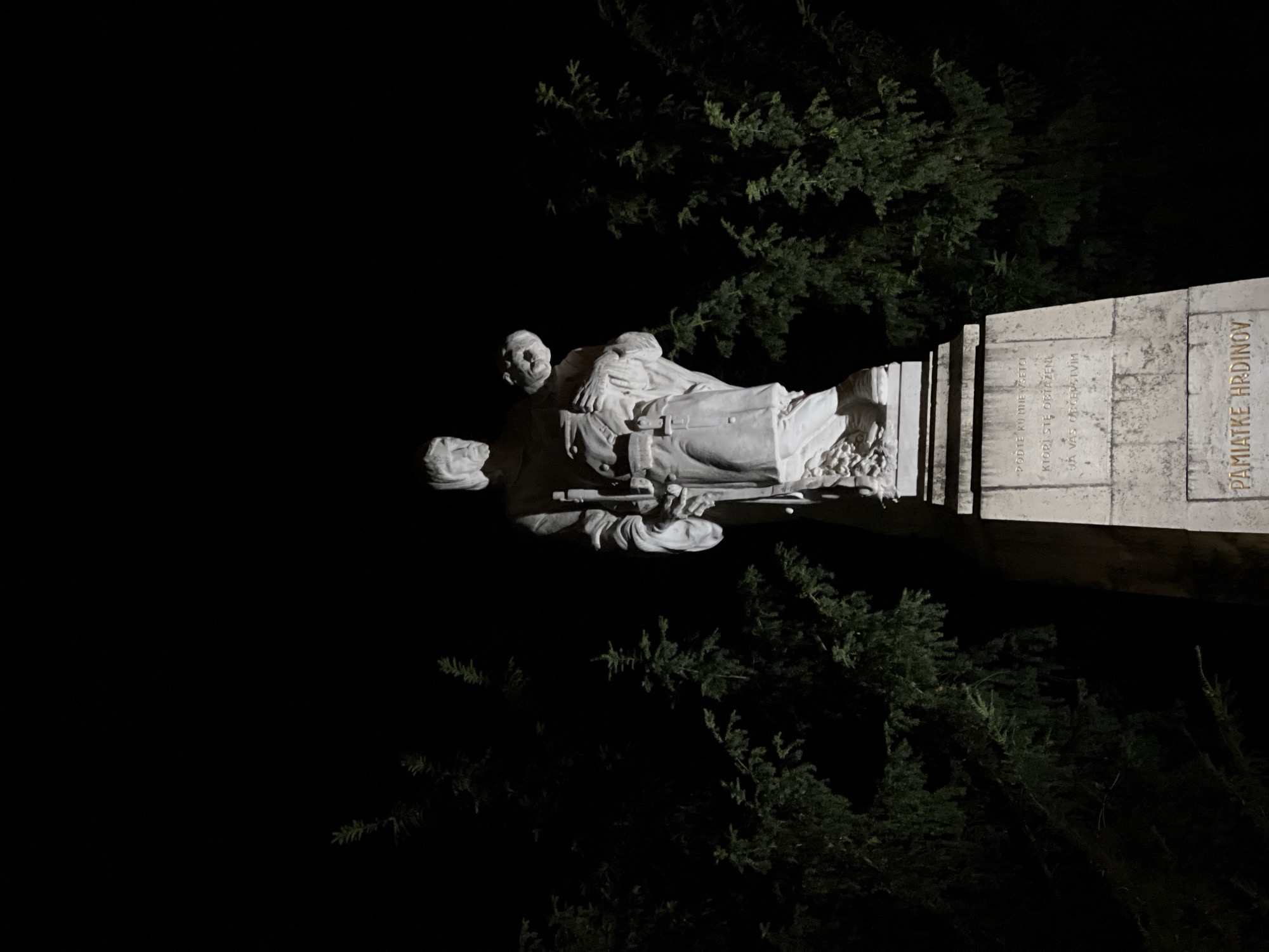 The image depicts a white stone sculpture of a man carrying a child at night, set against a dark background with evergreen trees on either side. The sculpture stands on a tiered stone pedestal with inscriptions. The inscription at the bottom of the pedestal reads "PAMIATKE HRDINOV," which translates to "Memorial to Heroes." Based on the inscription and visual context, this monument is likely located in Nitra, Slovakia, and is dedicated to fallen heroes, possibly during a conflict. The lighting suggests artificial illumination of the monument.