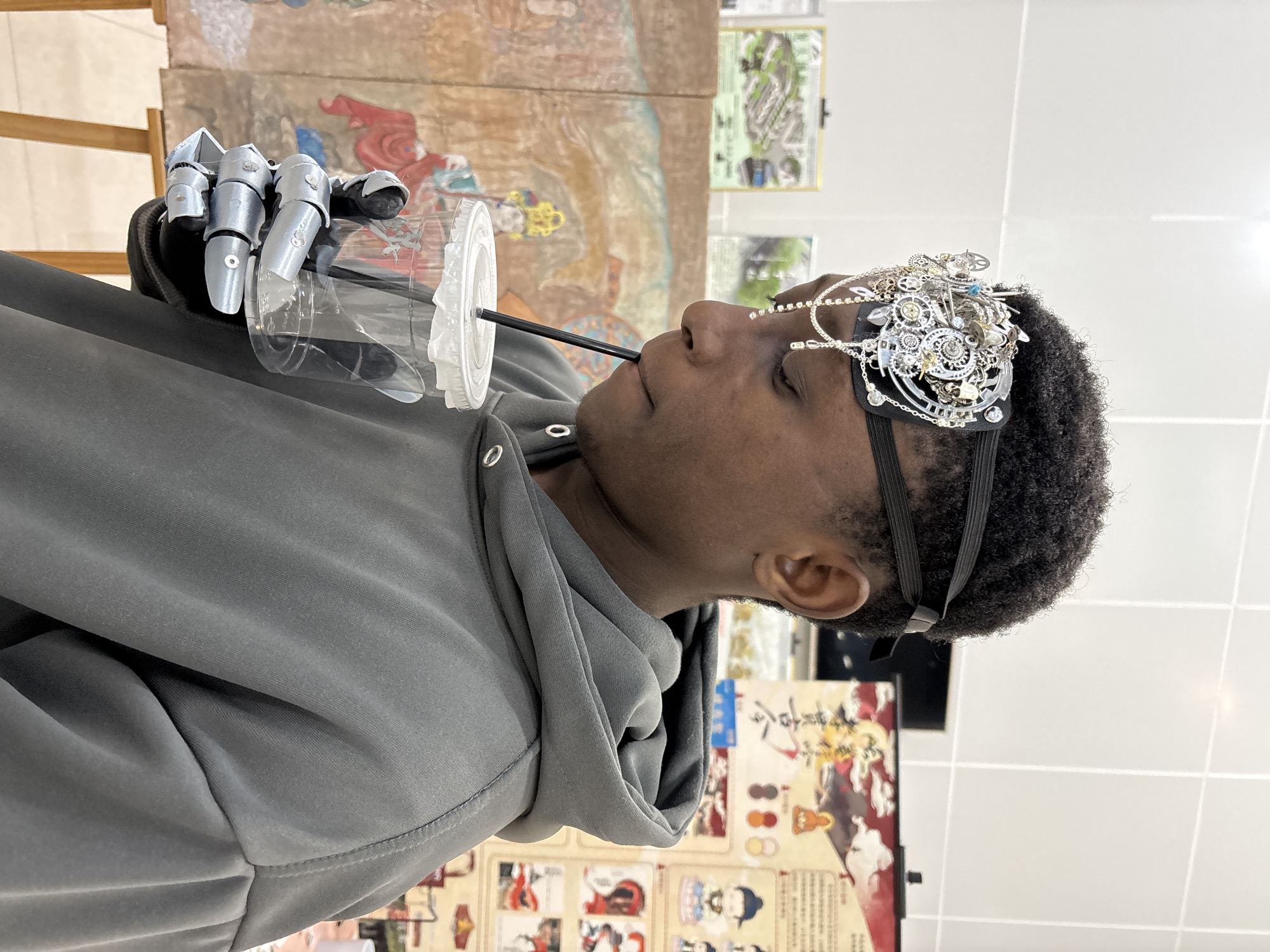A person with dark skin and short, curly hair is drinking from a clear plastic cup with a black straw. The person is wearing a gray hooded sweatshirt and an ornate, silver-colored headpiece with gears and other embellishments. The person's right hand is a robotic prosthetic. The person is indoors, with artwork and posters visible in the background. One piece of artwork appears to be a traditional Asian painting. The floor is tiled. The image was taken in Jiangjin, China.