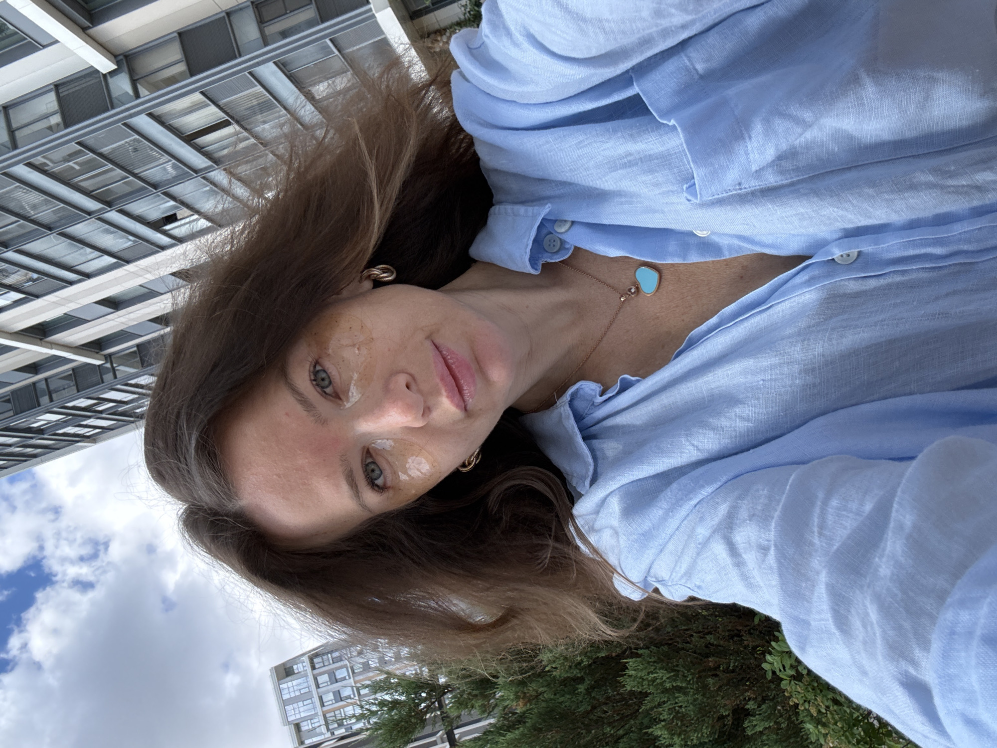 A woman with long brown hair is taking a selfie outdoors, with modern buildings and a cloudy sky in the background. She is wearing a light blue linen shirt and a necklace with a turquoise heart pendant. The woman has under-eye patches on. The location appears to be an urban environment with contemporary architecture, possibly in Moscow, Russia, given the style of the buildings.